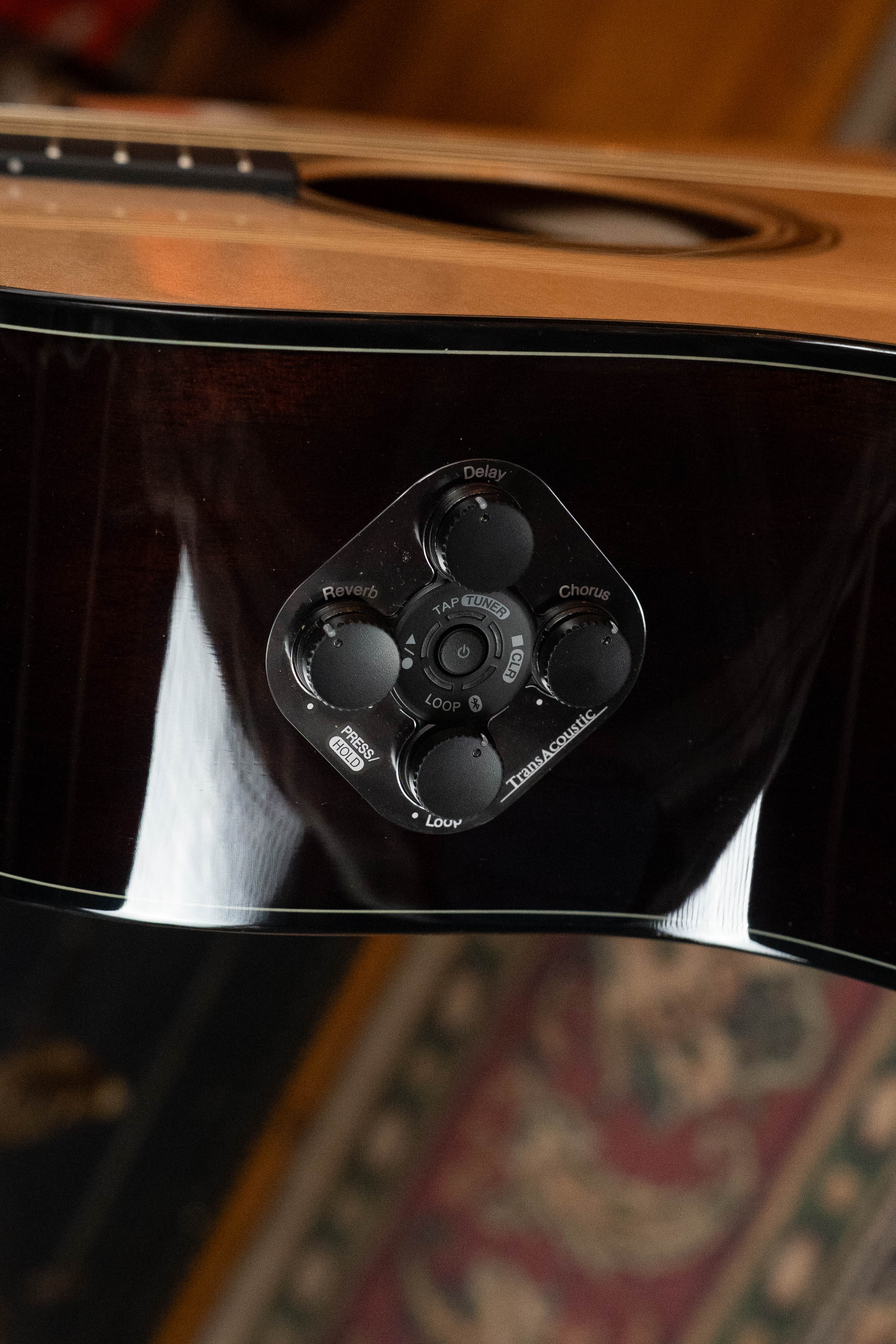 Close-up of the Yamaha TAG3C-NT Sitka/Mahogany Natural Dreadnought Guitar’s new TransAcoustic control panel with built-in reverb, delay, and chorus effects, shown on the guitar’s side with strings and a patterned rug in the background.