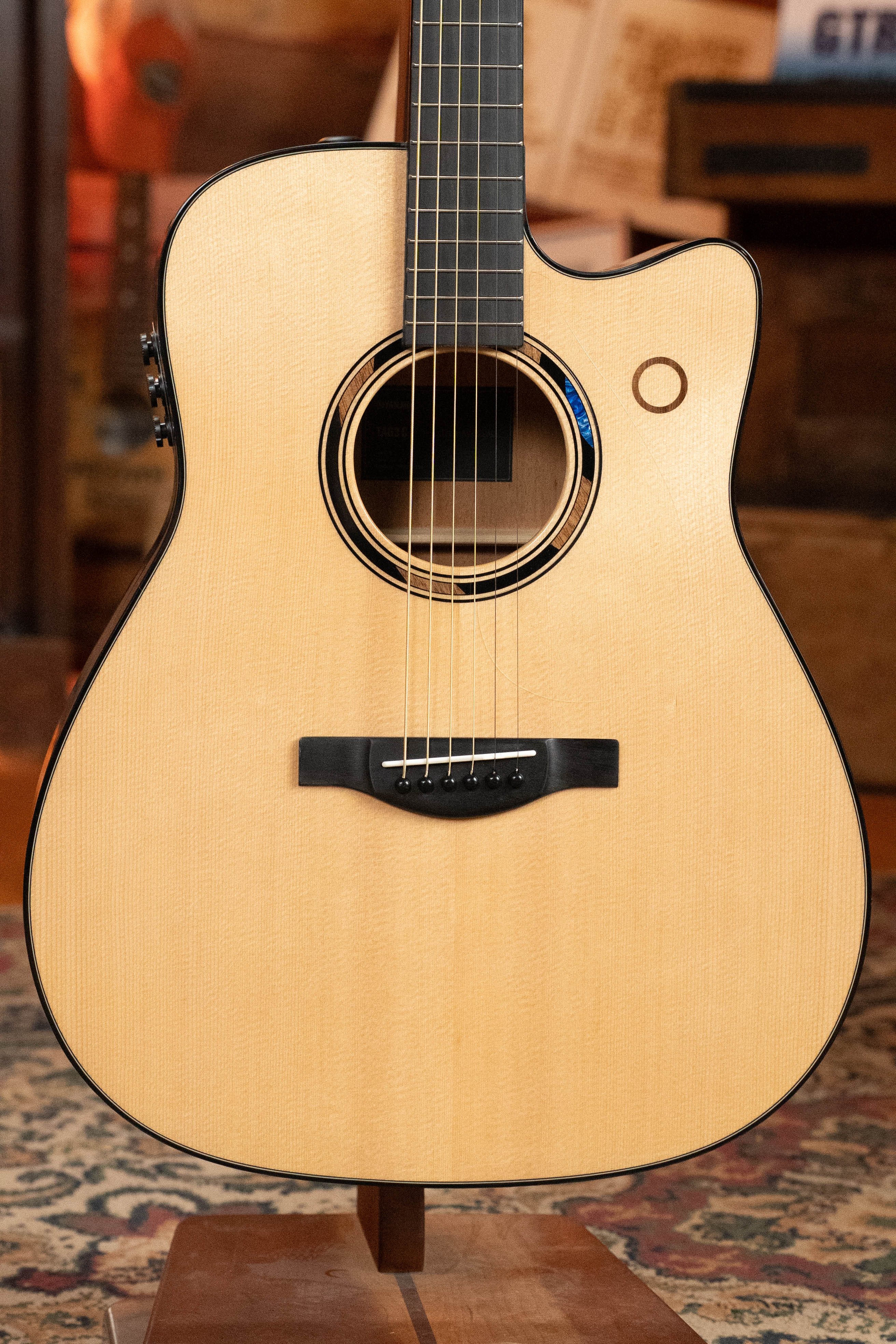 A close-up of the Yamaha TAG3C-NT Sitka/Mahogany Natural Dreadnought Guitar with the newest TransAcoustic System, featuring a light wood finish, black binding, and cutaway design—displayed upright against a warm indoor background.