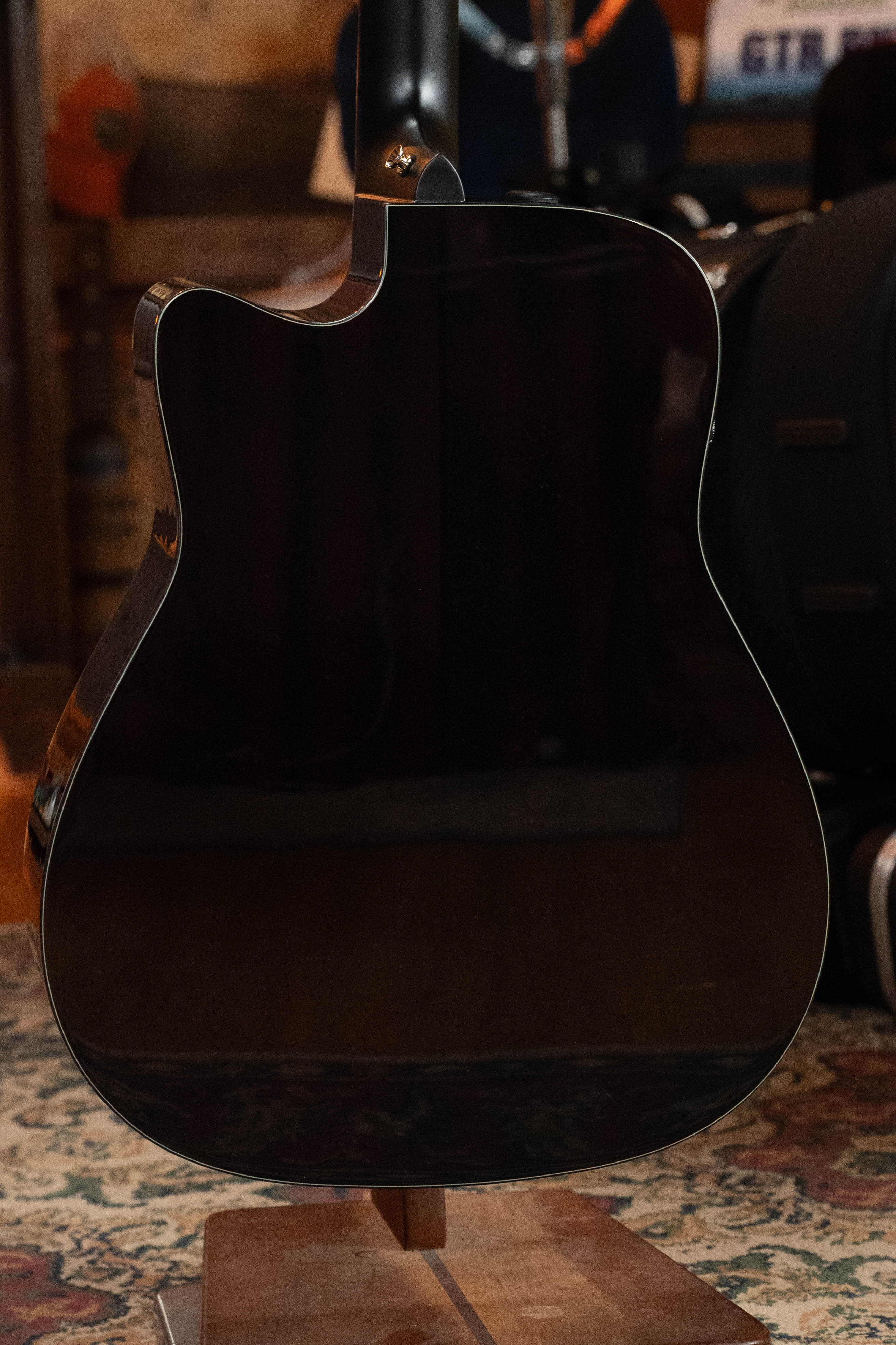 A close-up of the back of a Yamaha TAG3C-SDB Sitka/Mahogany Sandburst Dreadnought Guitar with the TransAcoustic System, resting on a stand with a patterned rug and blurred musical gear in the background.
