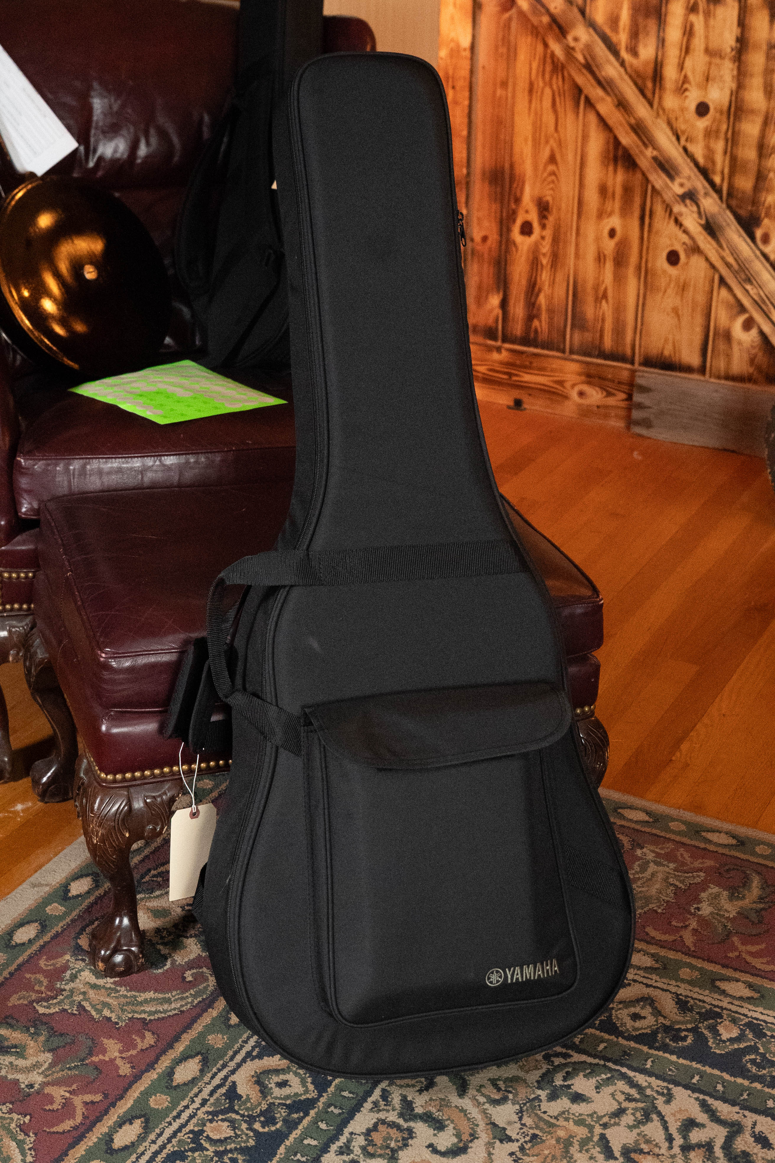 A Yamaha TAG3C-SDB Sitka/Mahogany Sandburst Dreadnought guitar with the newest TransAcoustic System (#0688) stands upright on a patterned rug next to a maroon leather chair in a warmly lit room with wooden walls and floors.