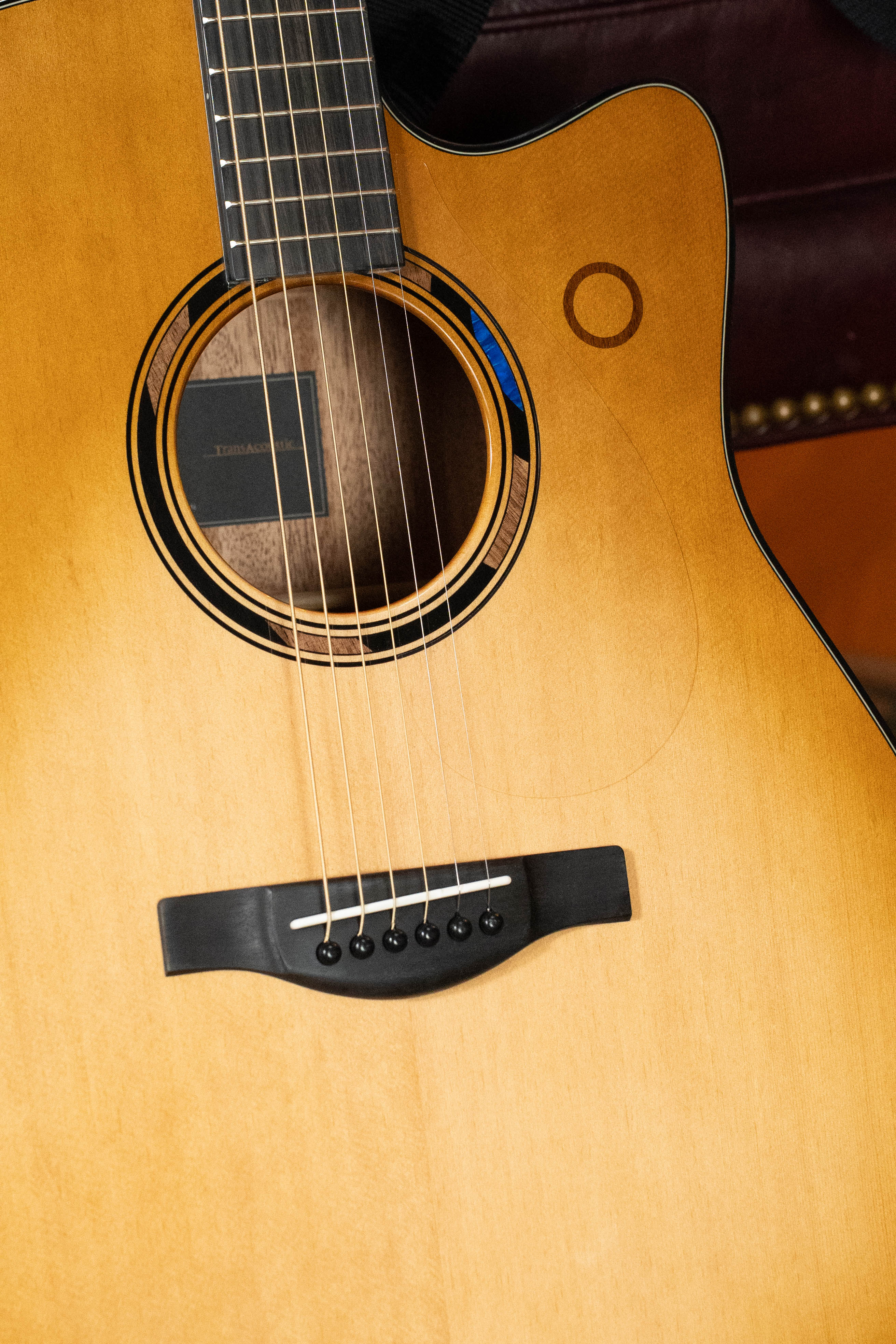 Close-up of a Yamaha TAG3C-SDB Sitka/Mahogany Sandburst Dreadnought Guitar, featuring strings, sound hole, and light wood. The NEWEST TransAcoustic System boosts its tone. A brown leather chair with brass studs is in the background.