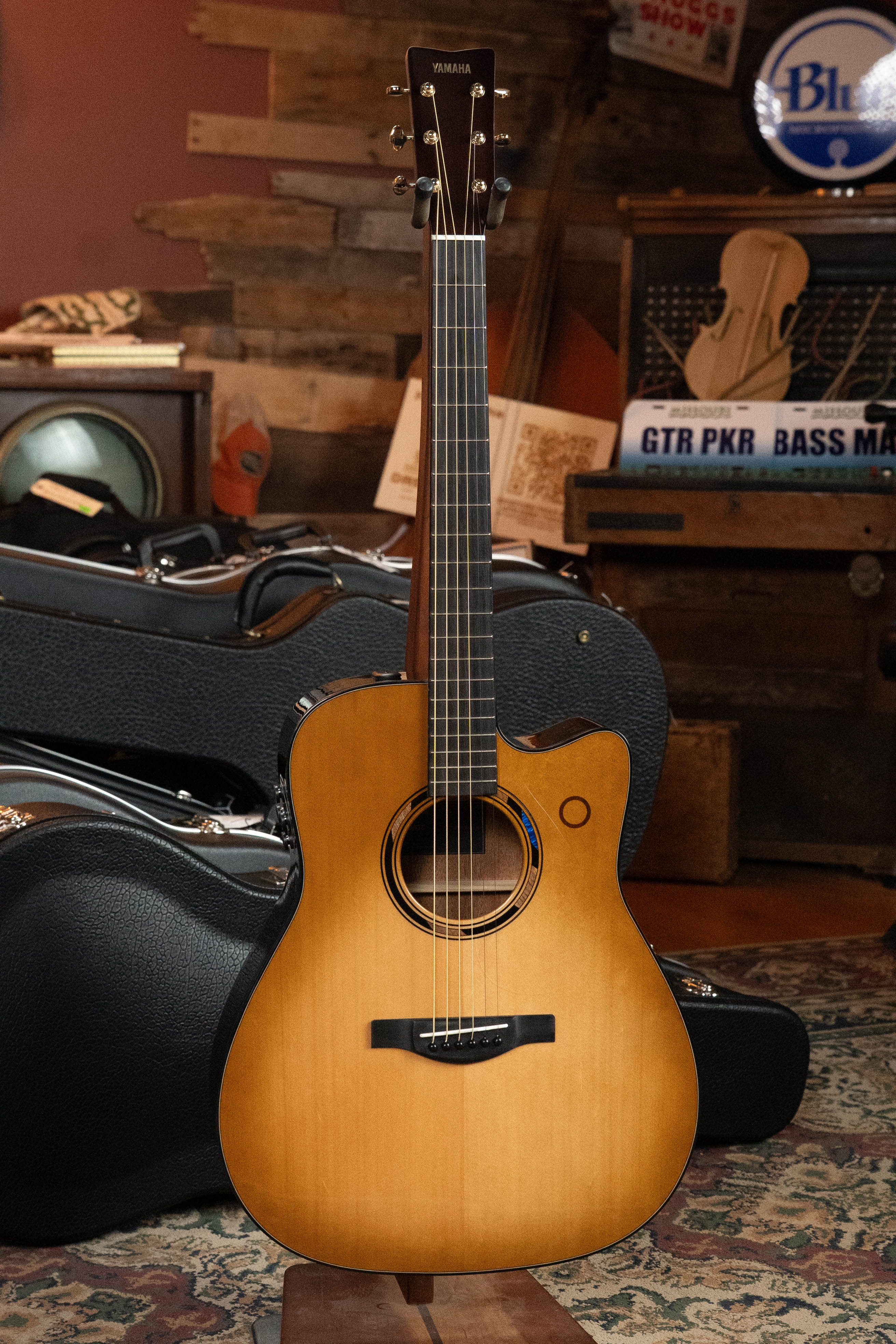 A Yamaha TAG3C-SDB Sitka/Mahogany Sandburst Dreadnought Guitar with the NEWEST TransAcoustic System #0688 stands upright in a cozy room, surrounded by instrument cases, music-inspired decor, and inviting ambient lighting.