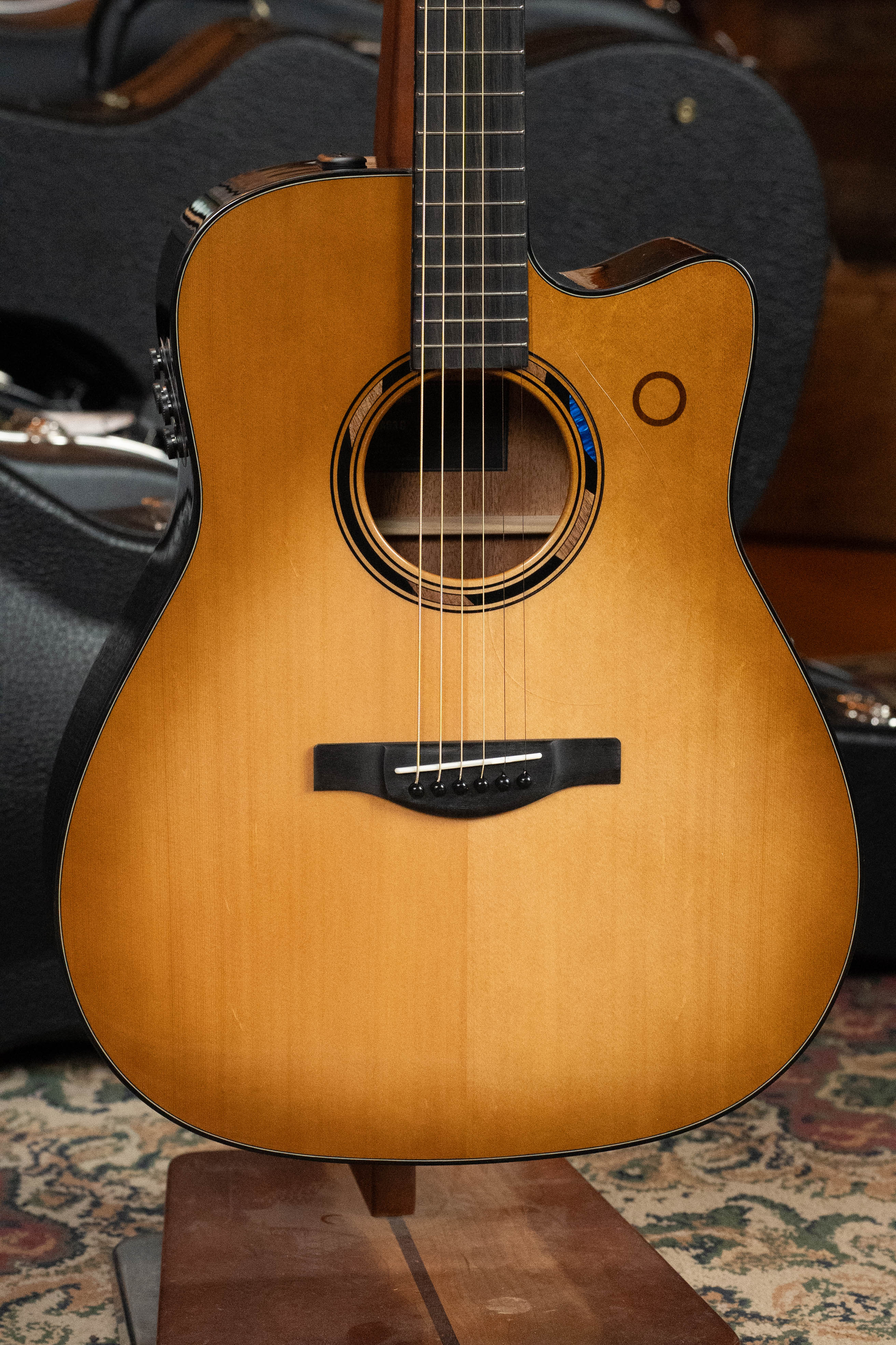 A Yamaha TAG3C-SDB Sitka/Mahogany Sandburst Dreadnought Guitar with the newest TransAcoustic System (#0688) rests on a stand atop a patterned rug, its black case in the background.