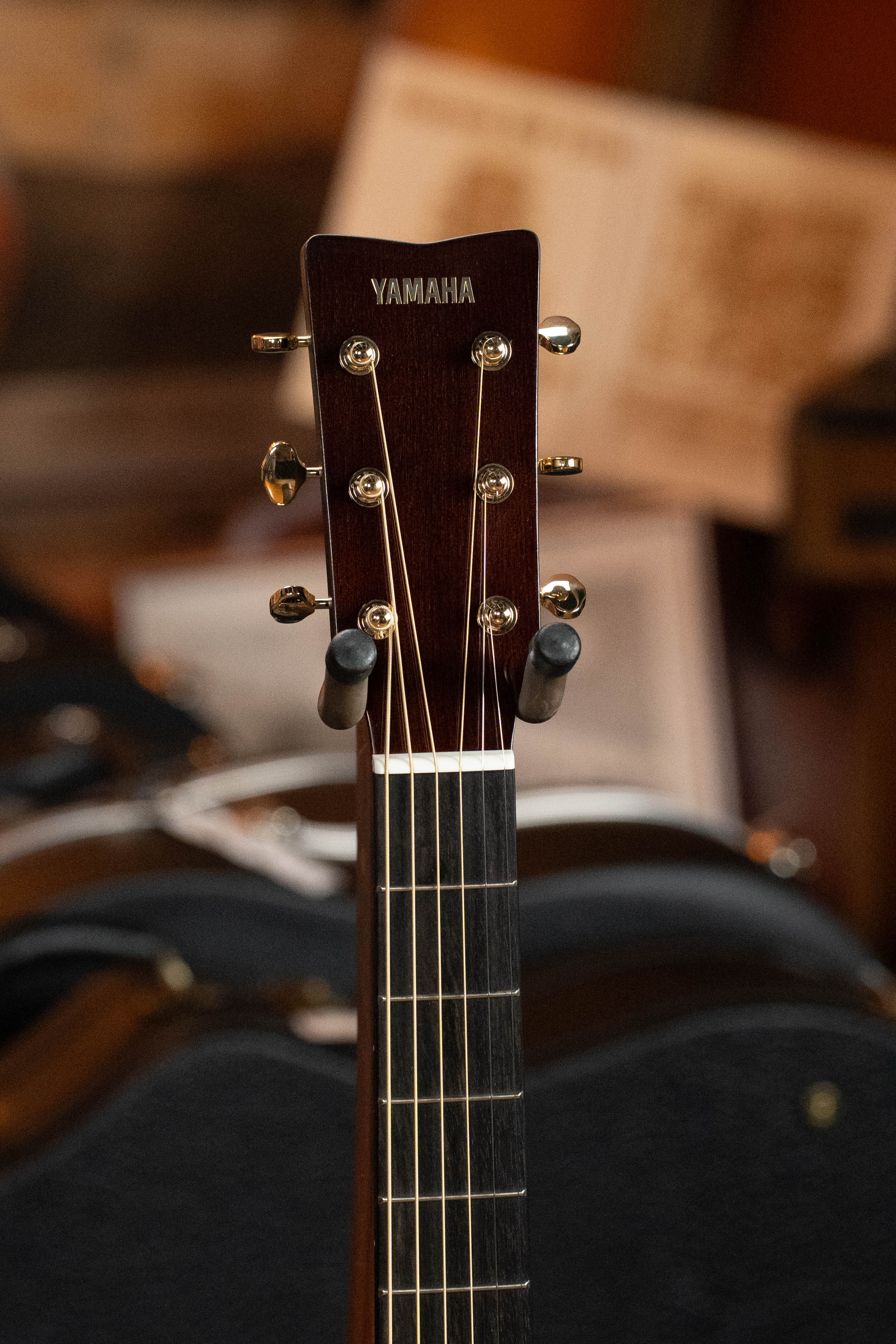 Close-up of the Yamaha TAG3C-SDB Sitka/Mahogany Sandburst Dreadnought headstock, showing tuning pegs and strings, with blurred guitars and sheet music behind, highlighting Yamaha’s NEWEST TransAcoustic System.