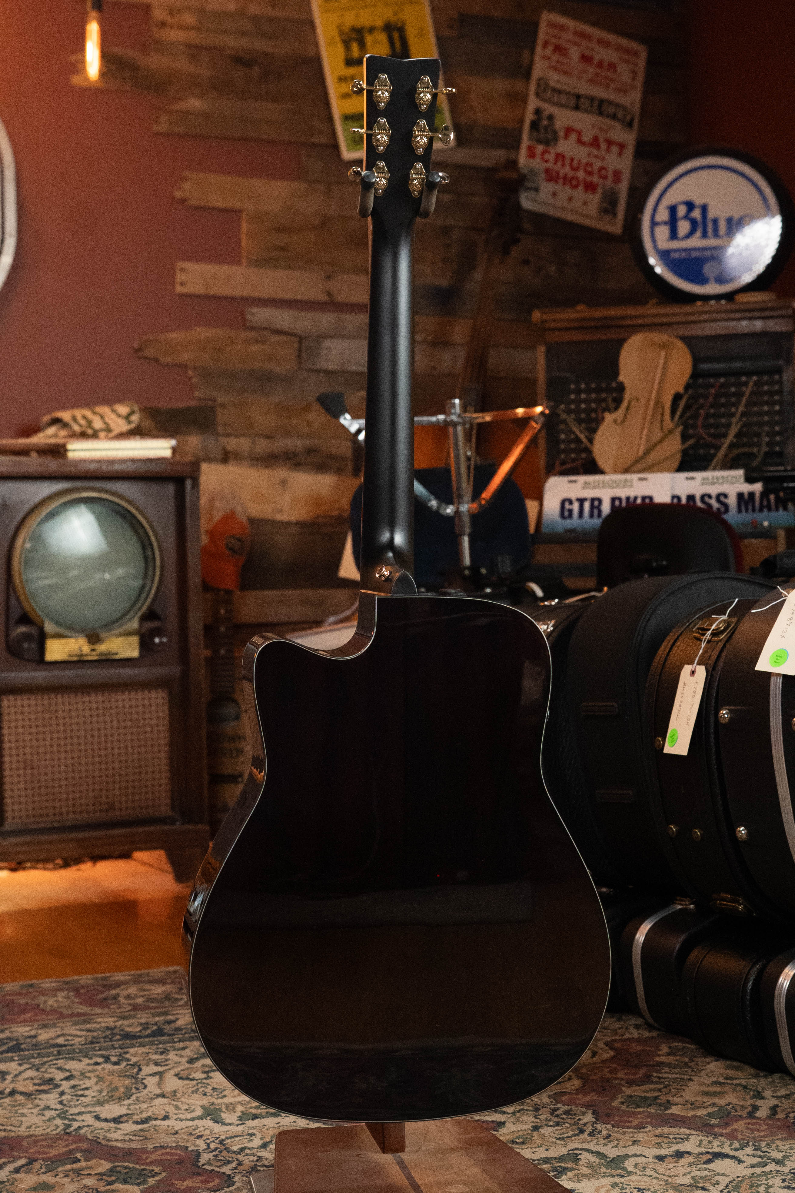 A Yamaha TAG3C-SDB Sitka/Mahogany Sandburst Dreadnought Guitar with the newest TransAcoustic System #0688 is displayed with its back facing the camera in a cozy, vintage-style room full of instruments, cases, a TV, and decor.