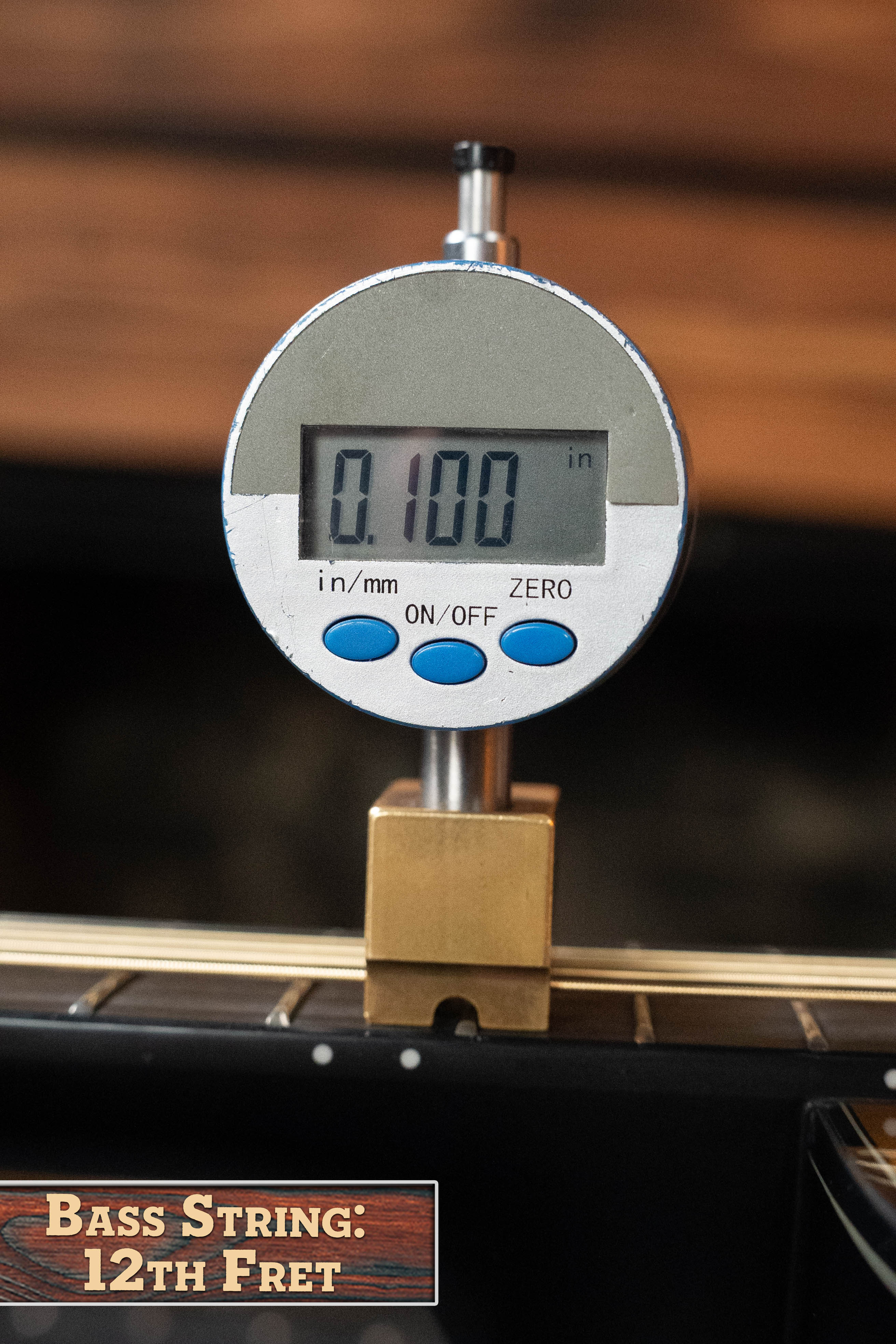 A digital gauge shows 0.100 in while measuring the height of a Yamaha TAG3C-SDB Sandburst Dreadnought guitar’s bass string above the 12th fret. A label at the bottom left reads Bass String: 12th Fret.