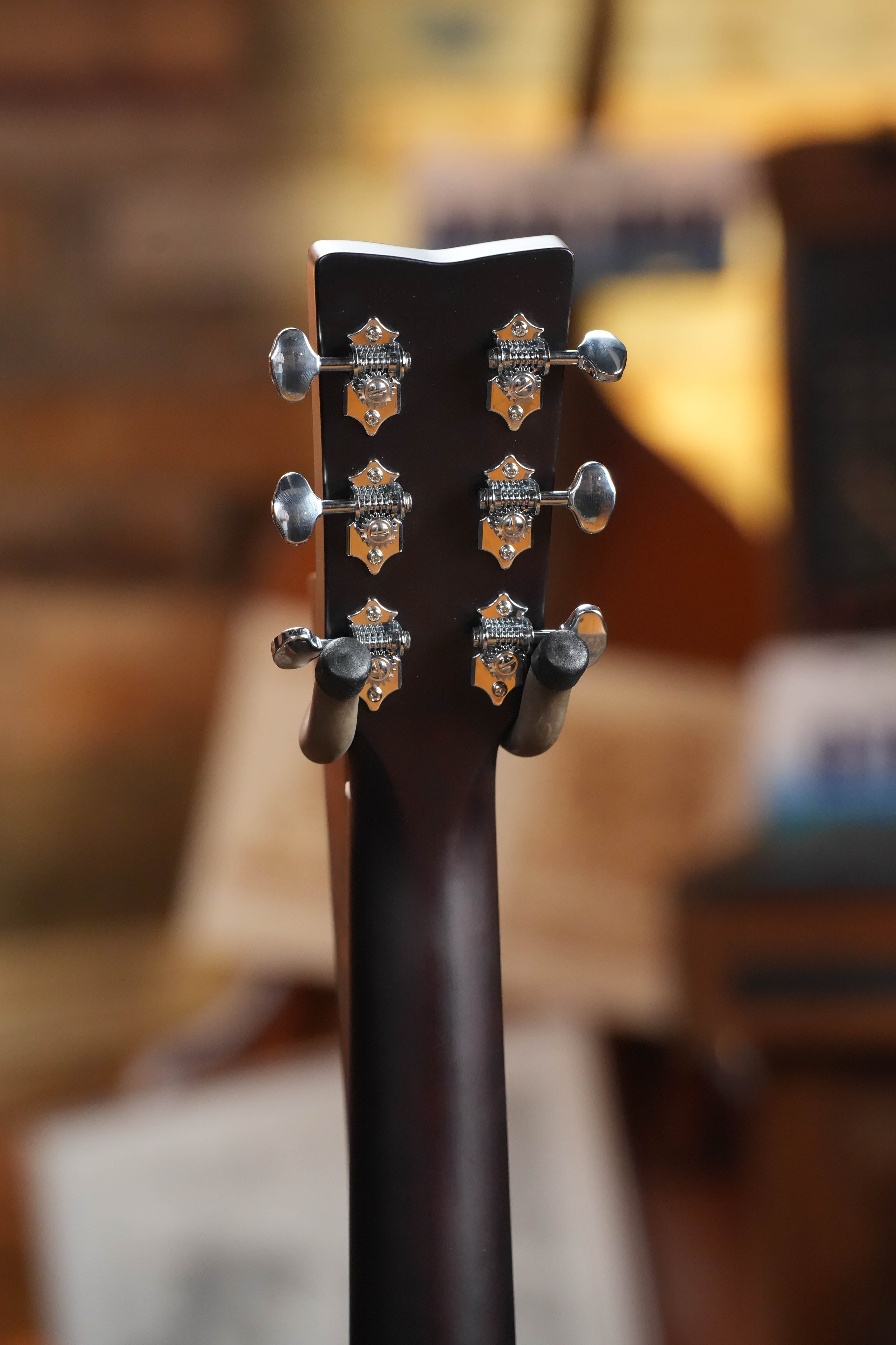 Close-up of the back of a Yamaha TAS1 C Sitka/Mahogany Sand Burst Transacoustic Concert Guitar headstock showing six tuning pegs, with a blurred background of warm-toned objects and papers.