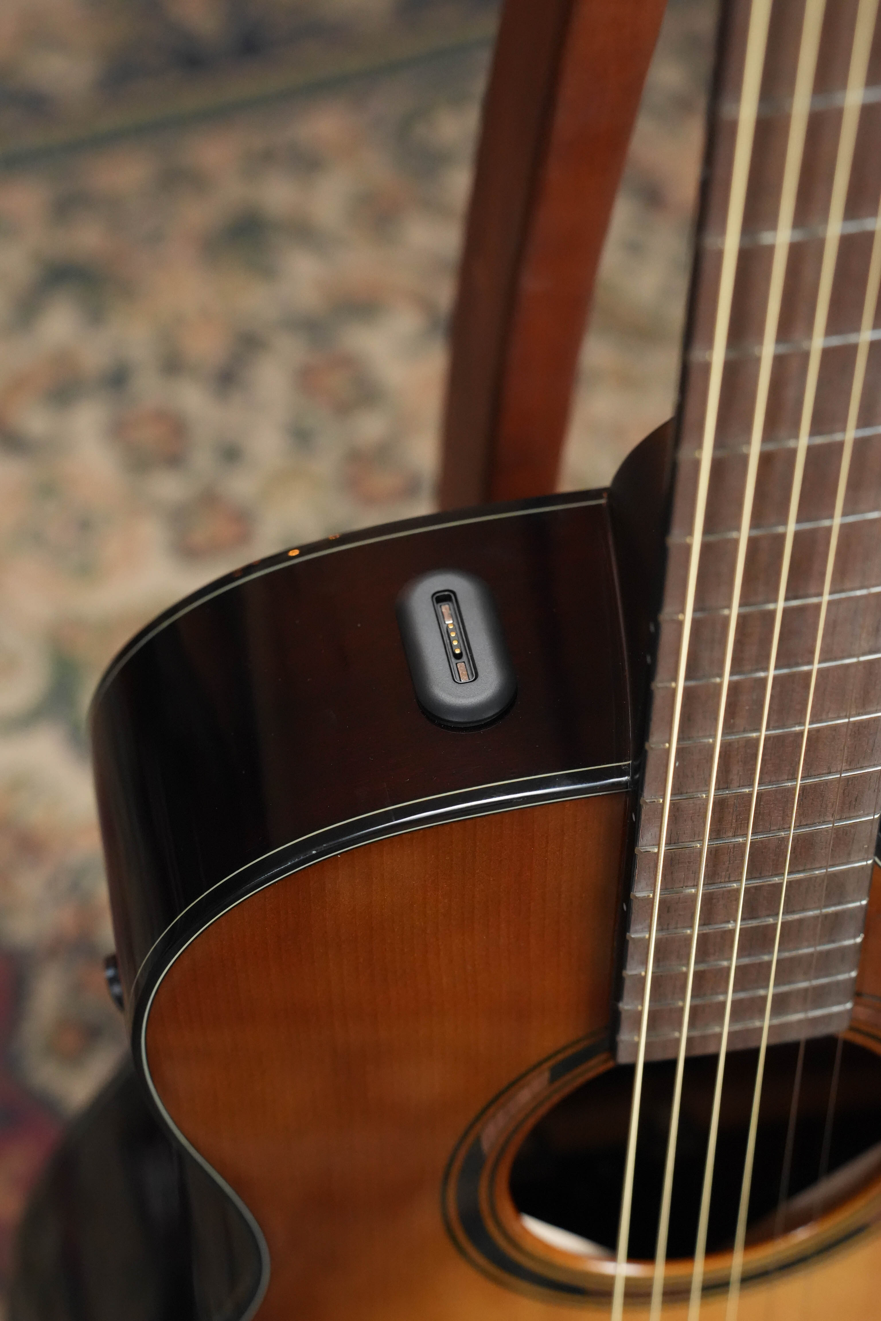 A close-up of the Yamaha TAS1 C Sitka/Mahogany Sand Burst Transacoustic Concert Guitar shows an electronic device by the neck, with a brown patterned rug and wooden chair in the background.
