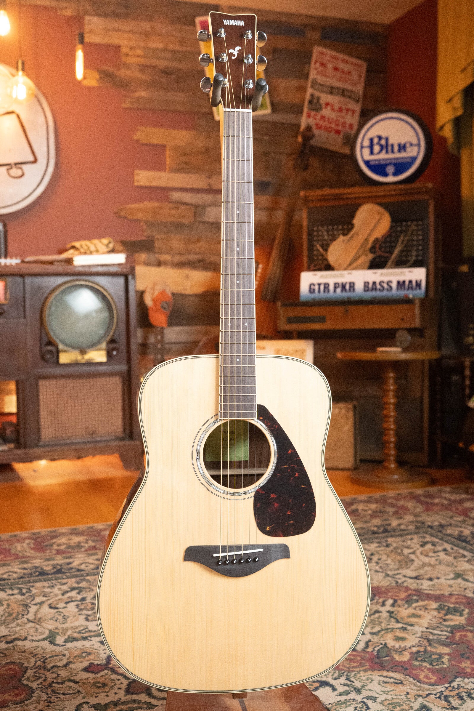 A Yamaha FG830 Spruce/Laminate Rosewood Natural Dreadnought Acoustic Guitar (#0562) by Yamaha stands upright on a patterned rug in a cozy, vintage-style room with warm lighting and retro décor.