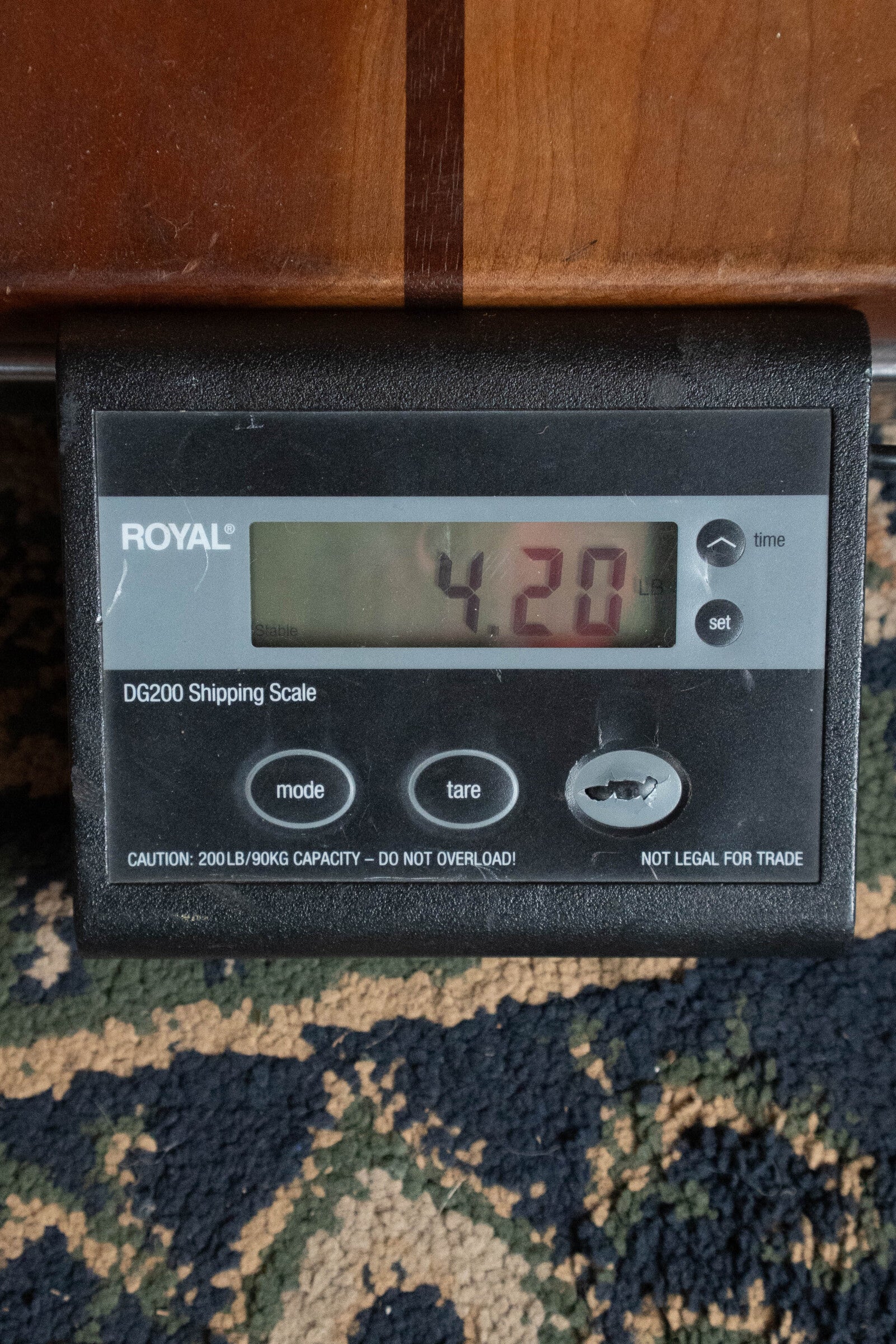 A digital scale on a patterned carpet shows 4.20 pounds, enough to weigh items like the Yamaha FG830 Spruce/Laminate Rosewood Natural Dreadnought Acoustic Guitar by Yamaha. Part of a wooden surface appears above the black scale.
