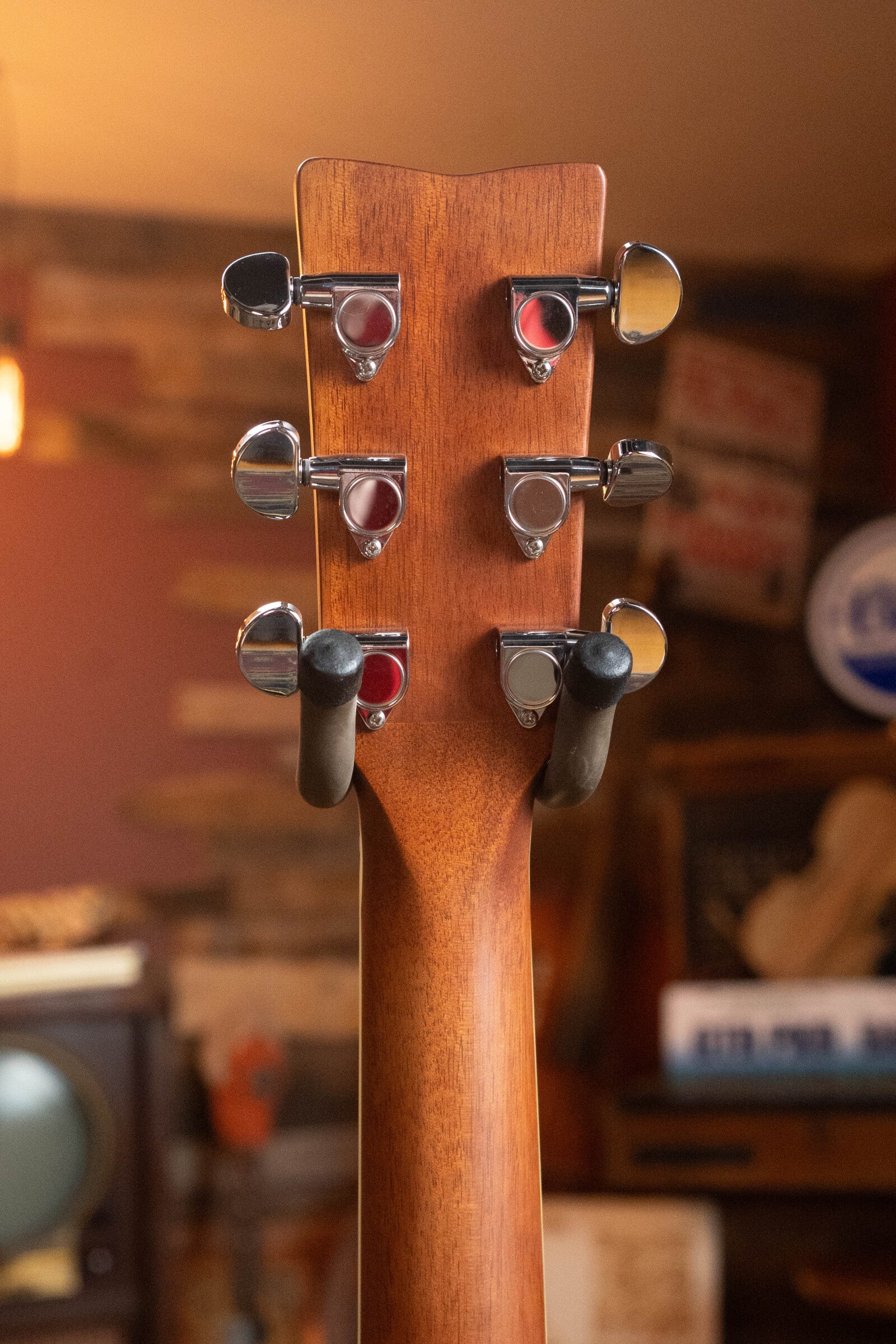 Close-up of the Yamaha FG830 Spruce/Laminate Rosewood Natural Dreadnought Acoustic Guitar #0600 headstock with six tuning pegs, set against a cozy room with warm lighting, wood-paneled walls, vintage signs, and a retro TV.