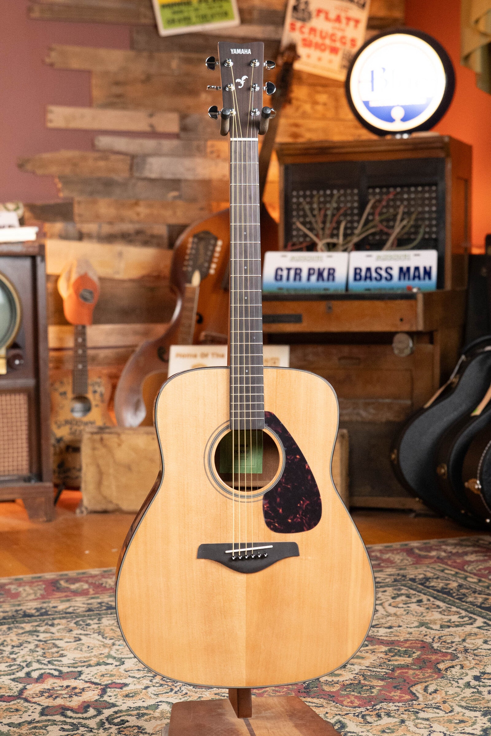 A Yamaha GigMaker Deluxe Acoustic Pack - Natural Finish #8229 by Yamaha is displayed upright on a stand in a cozy, vintage-style room filled with musical instruments and decorative signs.
