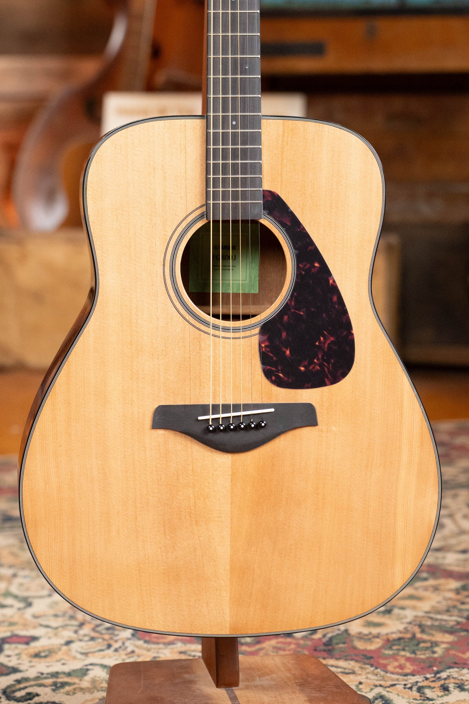 A Yamaha GigMaker Deluxe Acoustic Pack - Natural Finish #8229 dreadnought guitar with a black pickguard and bridge stands on a patterned rug with wooden furniture in the background—perfect for beginner musicians.