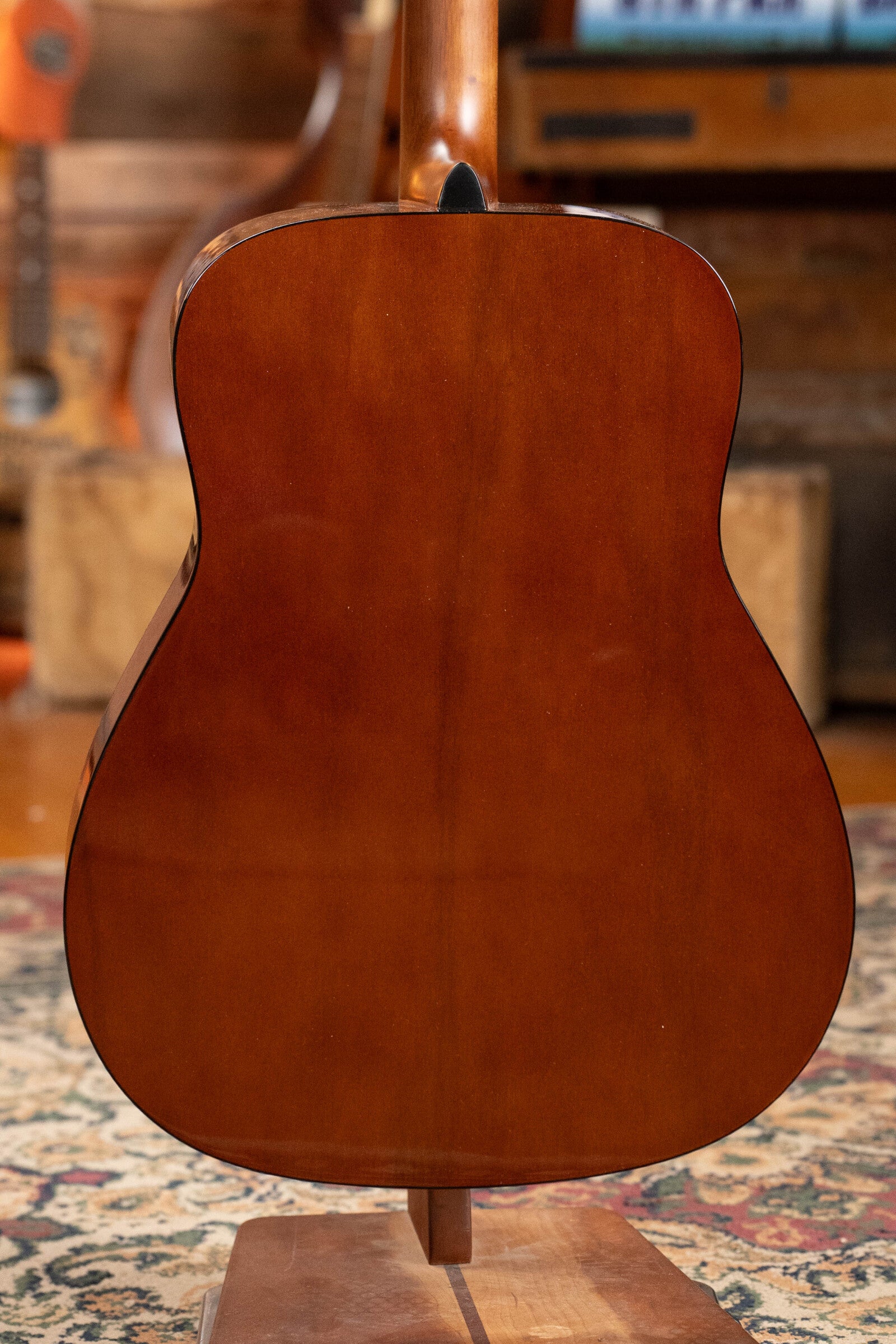 A Yamaha GigMaker Deluxe Acoustic Pack - Natural Finish #8229 by Yamaha stands upright on a wooden stand, showcasing its glossy brown back. A patterned rug and blurred wooden furniture are visible in the background.