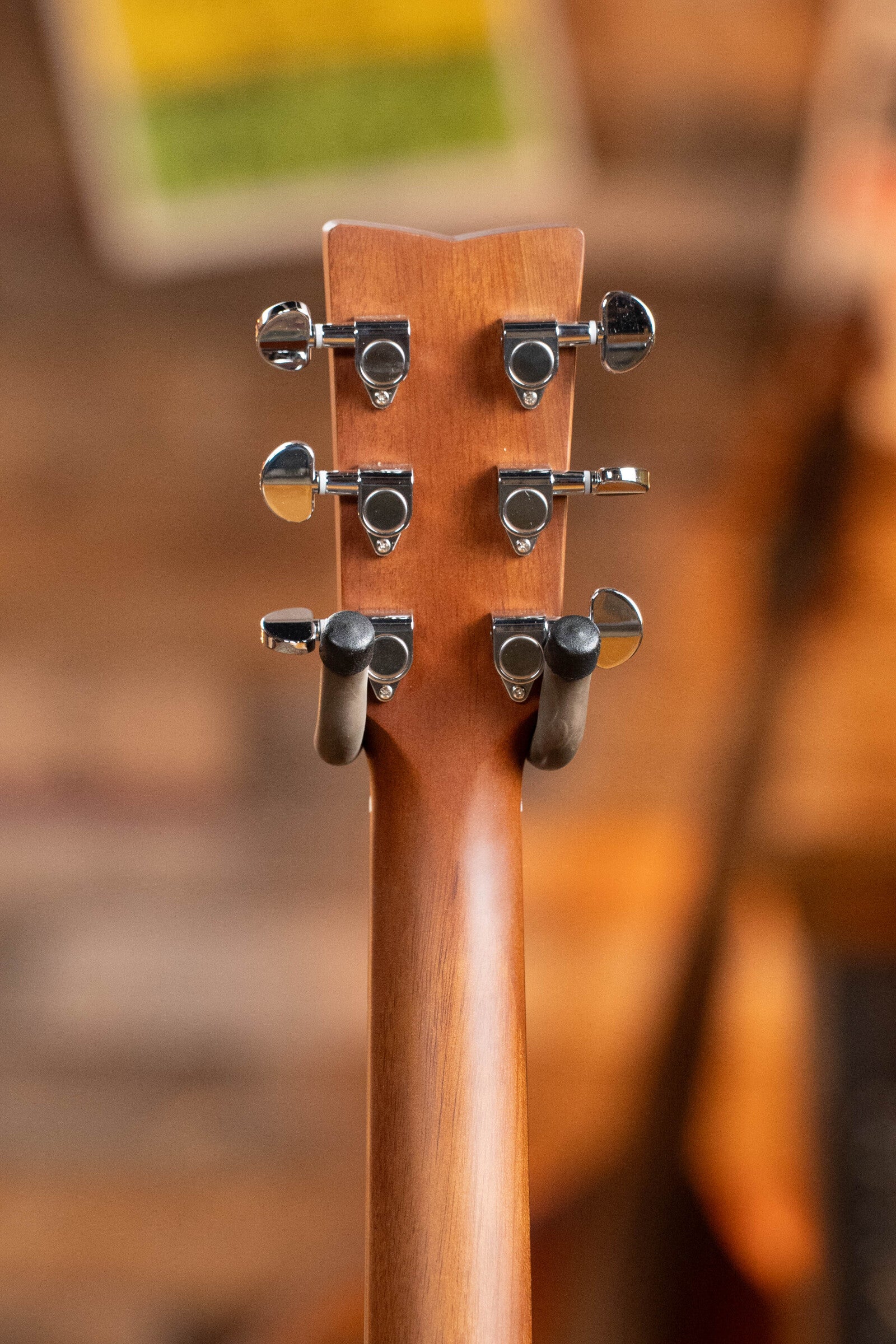 Close-up of the back of a Yamaha GigMaker Deluxe Acoustic Pack - Natural Finish #8229 headstock with six chrome tuning pegs, resting on a stand against a blurred wooden background and hints of colorful posters. Brand: Yamaha.