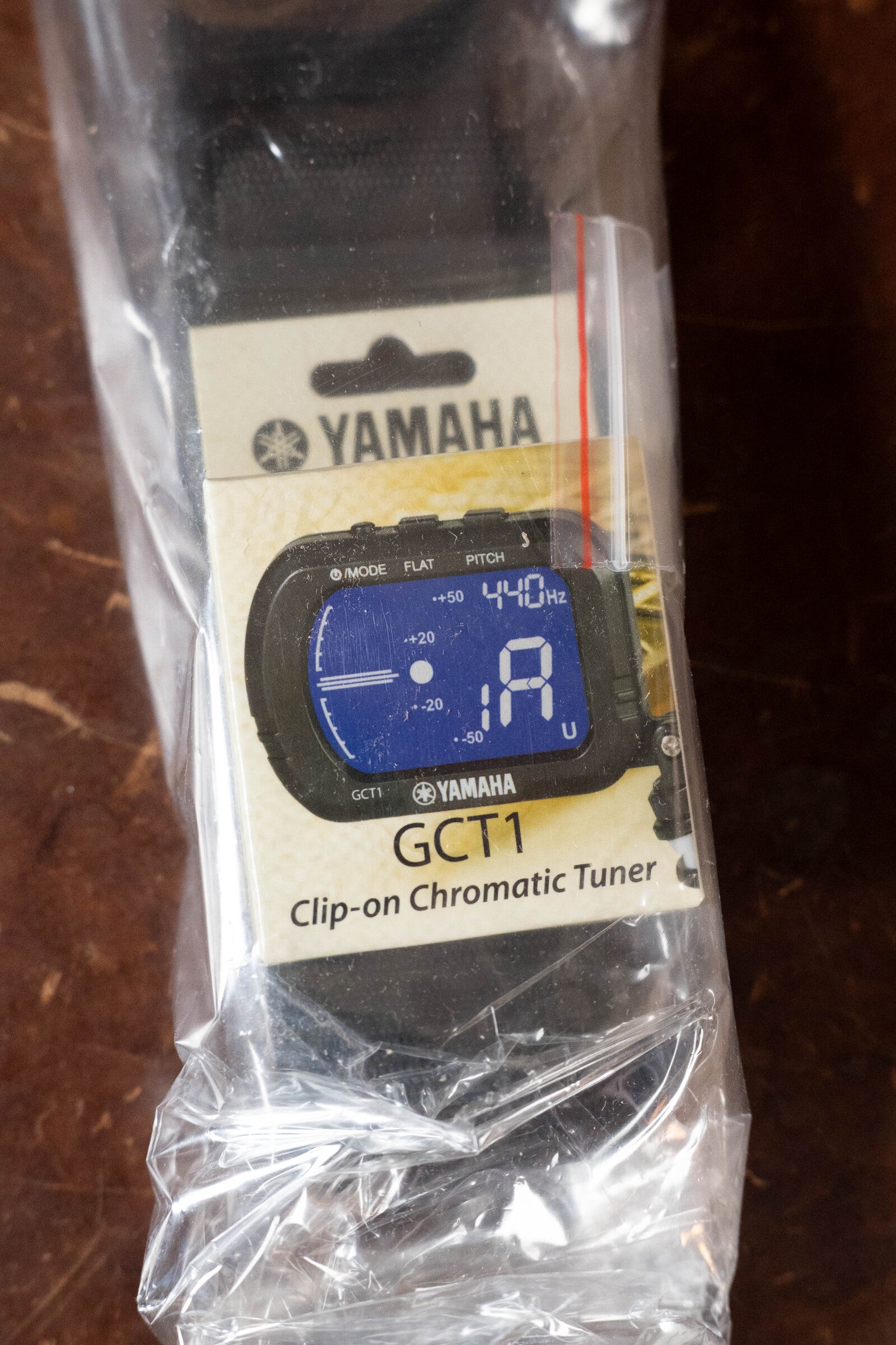 The Yamaha GCT1 clip-on chromatic tuner is displayed in its packaging, featuring a blue digital screen. Perfect for use with the Yamaha GigMaker Deluxe Acoustic Pack - Natural Finish #8229, it comes partially wrapped in clear plastic.