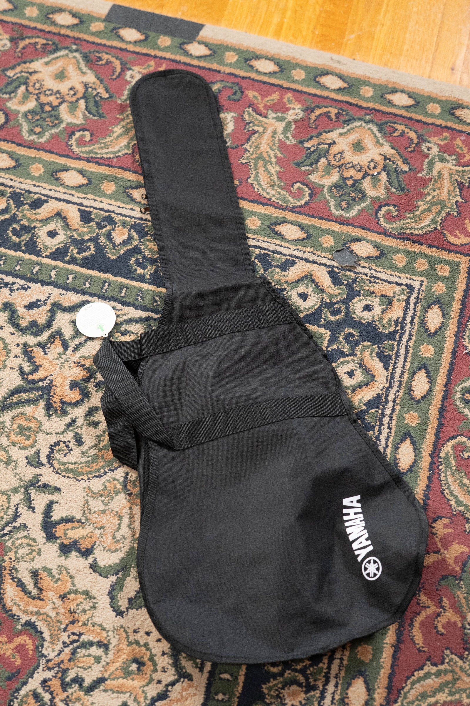 A black Yamaha gig bag from the Yamaha GigMaker Deluxe Acoustic Pack - Natural Finish #8229 rests on a patterned rug, with a wooden floor in the corner—ideal for your dreadnought acoustic guitar.