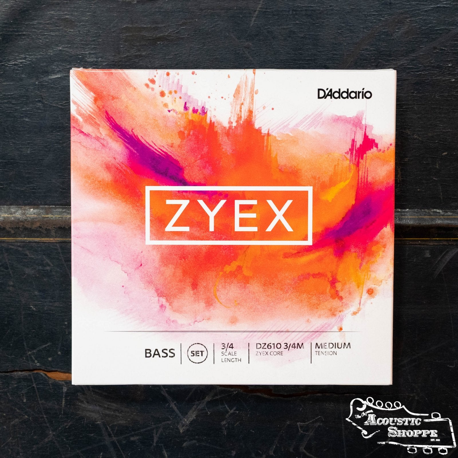 A box of D'Addario DZ610 3/4M Zyex Bass Strings with a bold orange and pink splash design rests on dark wood. Known for gut-like tone and warm pizzicato, the Acoustic Shoppe logo is displayed in the bottom right corner.