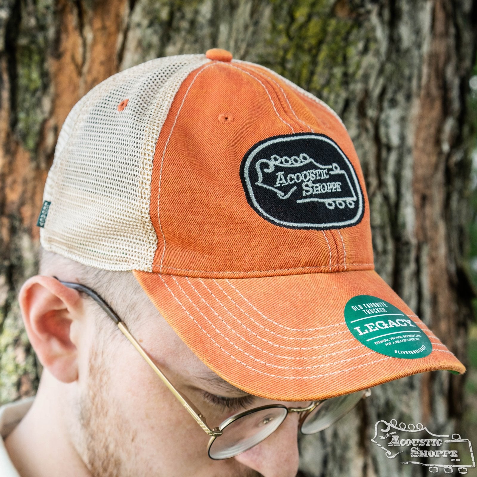 A person wearing glasses and an orange/black Acoustic Shoppe Legacy Badge Trucker Cap by Amplify Screen Printing & Promotions, featuring a green Legacy sticker, stands in front of a tree at this popular Eastman dealer.