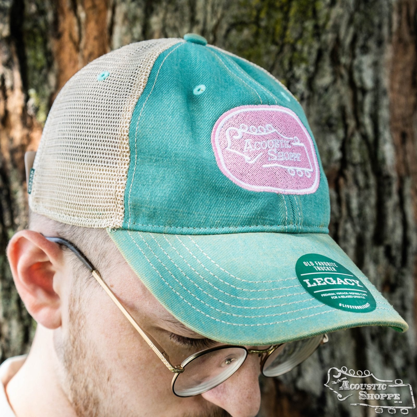 A person wearing glasses and an Acoustic Shoppe Legacy Badge Trucker Cap Teal / Pink by Amplify Screen Printing & Promotions, featuring a pink logo and eye-catching LEGACY sticker, stands in front of a tree.