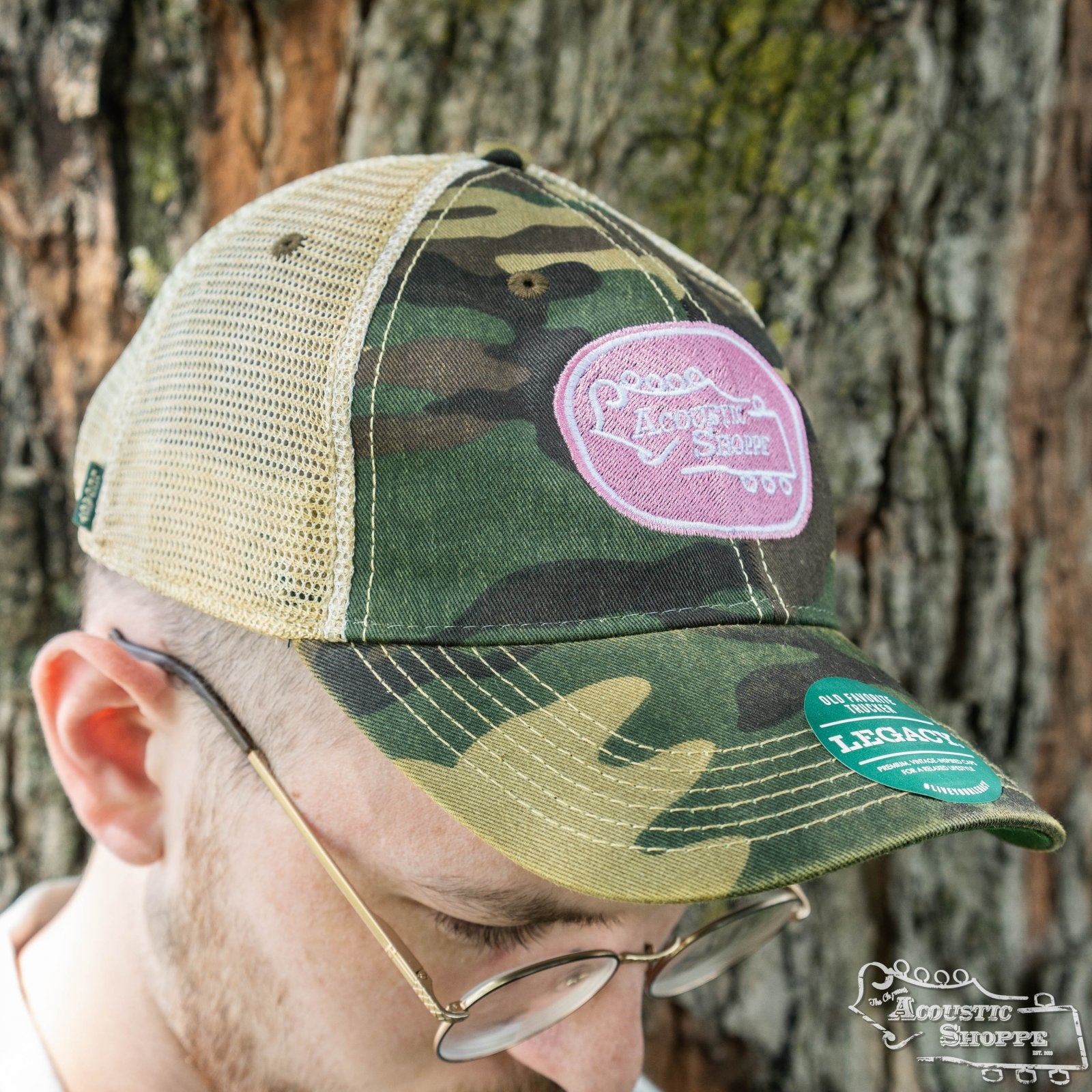 A person wearing glasses and an Amplify Screen Printing & Promotions Acoustic Shoppe Legacy Badge Trucker Cap Camo / Pink stands in front of a tree trunk.