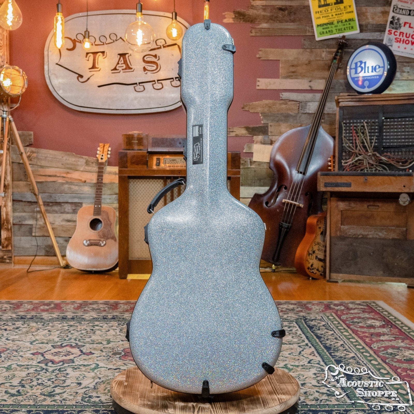 A sparkling Calton Cases Dreadnought D-18 Diamond Smooth Glitter Exterior/Burgundy Interior #21-1800007 stands on a wooden stand in a cozy music studio, surrounded by guitars, vintage gear, and warm lighting—ideal for the Acoustic Shoppe.