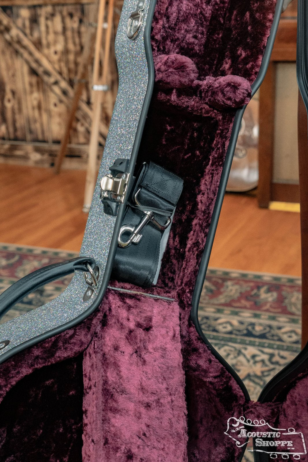 A close-up of the Calton Dreadnought D-18 Diamond Smooth Glitter case by Calton Cases, lined with plush burgundy fabric and holding a black leather strap, sits on a wooden floor with a patterned rug in a rustic room.