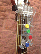 A close-up of a guitar neck fitted with the ChordBuddy Guitar Learning System (HL00123875) by Hal Leonard Corp., featuring colored buttons, highlights this innovative learning device against a textured brick wall background.