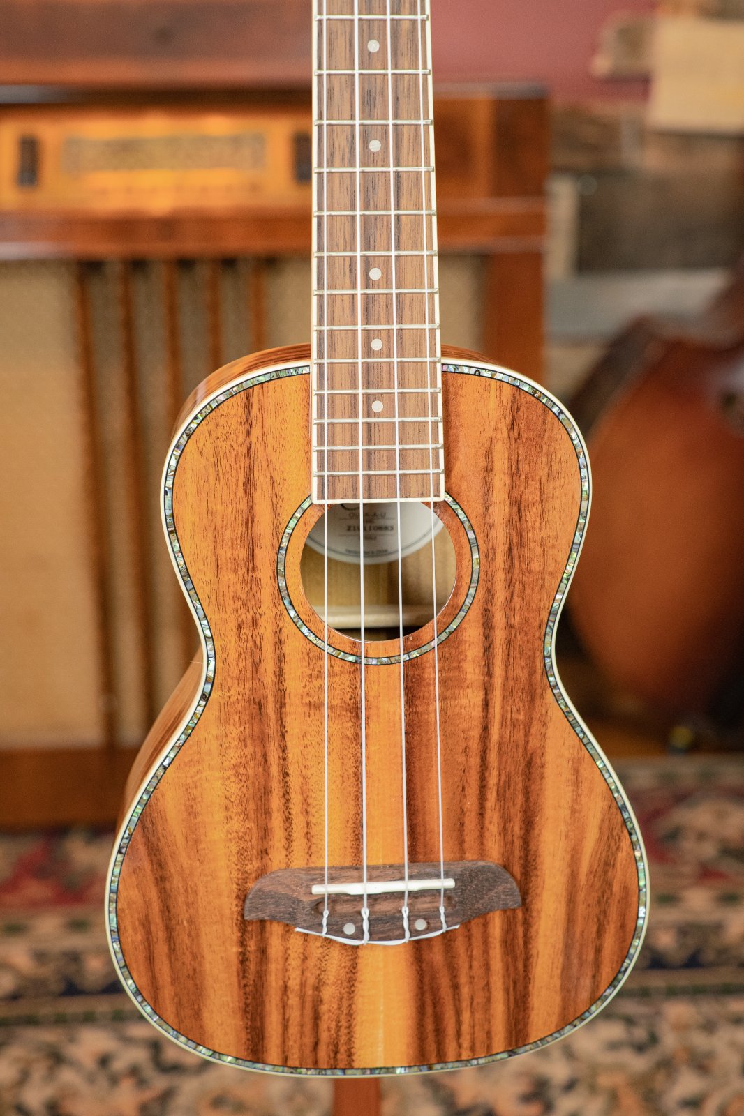 A close-up of the Oscar Schmidt OU5K-A-U KOA Professional Concert Ukulele, featuring detailed koa wood grain, abalone inlay, and four strings, is shown in front of a vintage radio on a patterned rug.