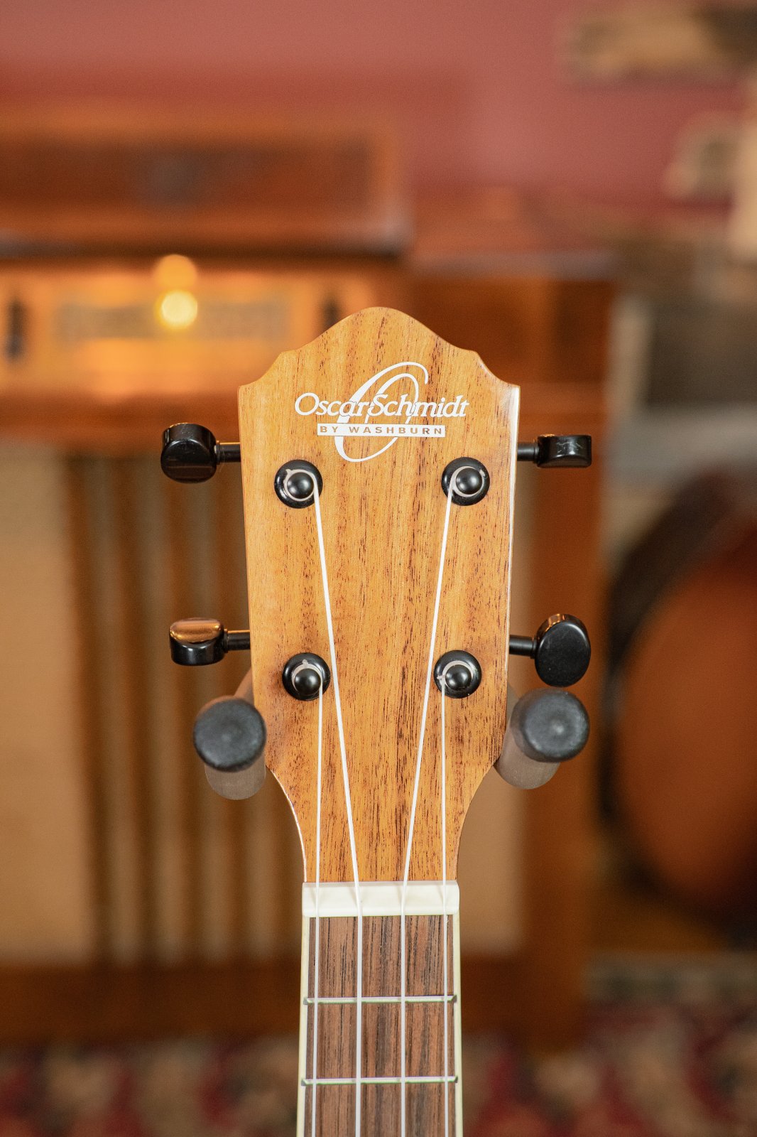 Close-up of the headstock of the Oscar Schmidt OU5K-A-U KOA Professional Concert Ukulele, highlighting tuning pegs and logo, with a blurred background in warm wooden tones and subtle hints of other musical instruments.