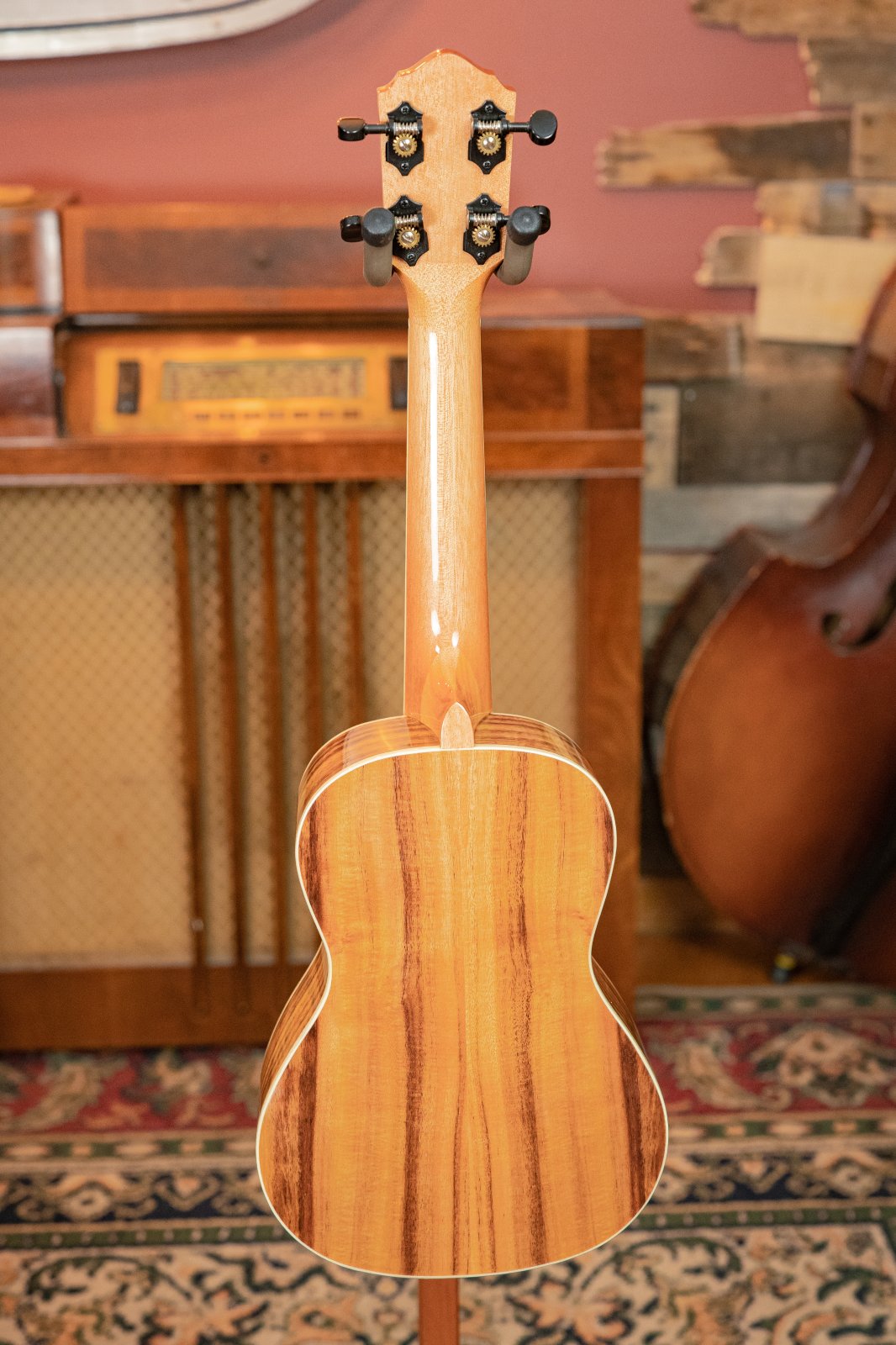 The Oscar Schmidt OU5K-A-U KOA Professional Concert Ukulele stands upright, highlighting its glossy Hawaiian Koa wood grain. In the background are a vintage radio, an upright bass, and a patterned rug.