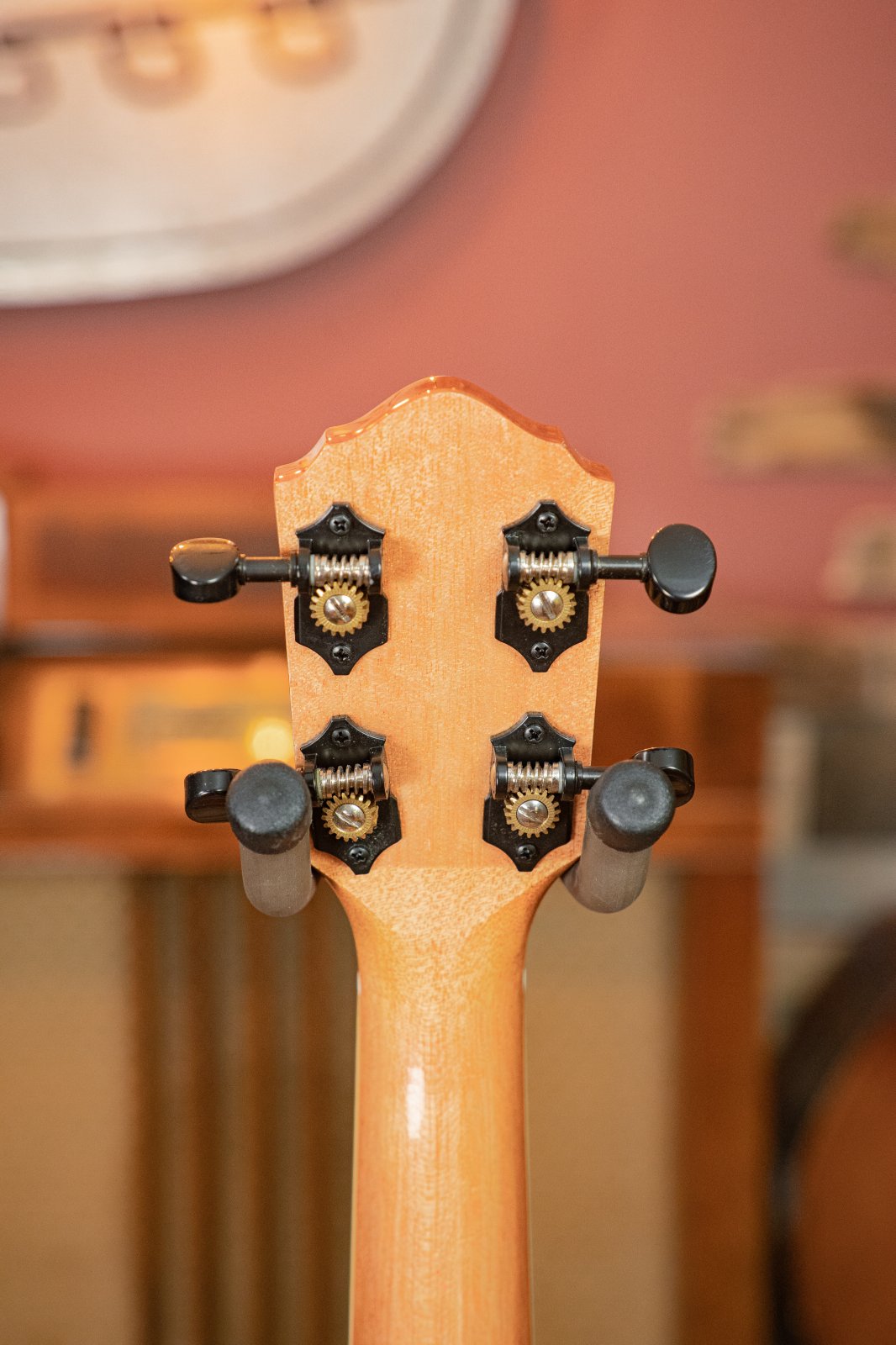 Close-up of the Oscar Schmidt OU5K-A-U KOA Professional Concert Ukulele’s headstock, featuring four black tuning pegs and metal gears, set against a blurred background of warm-toned furniture and decor.