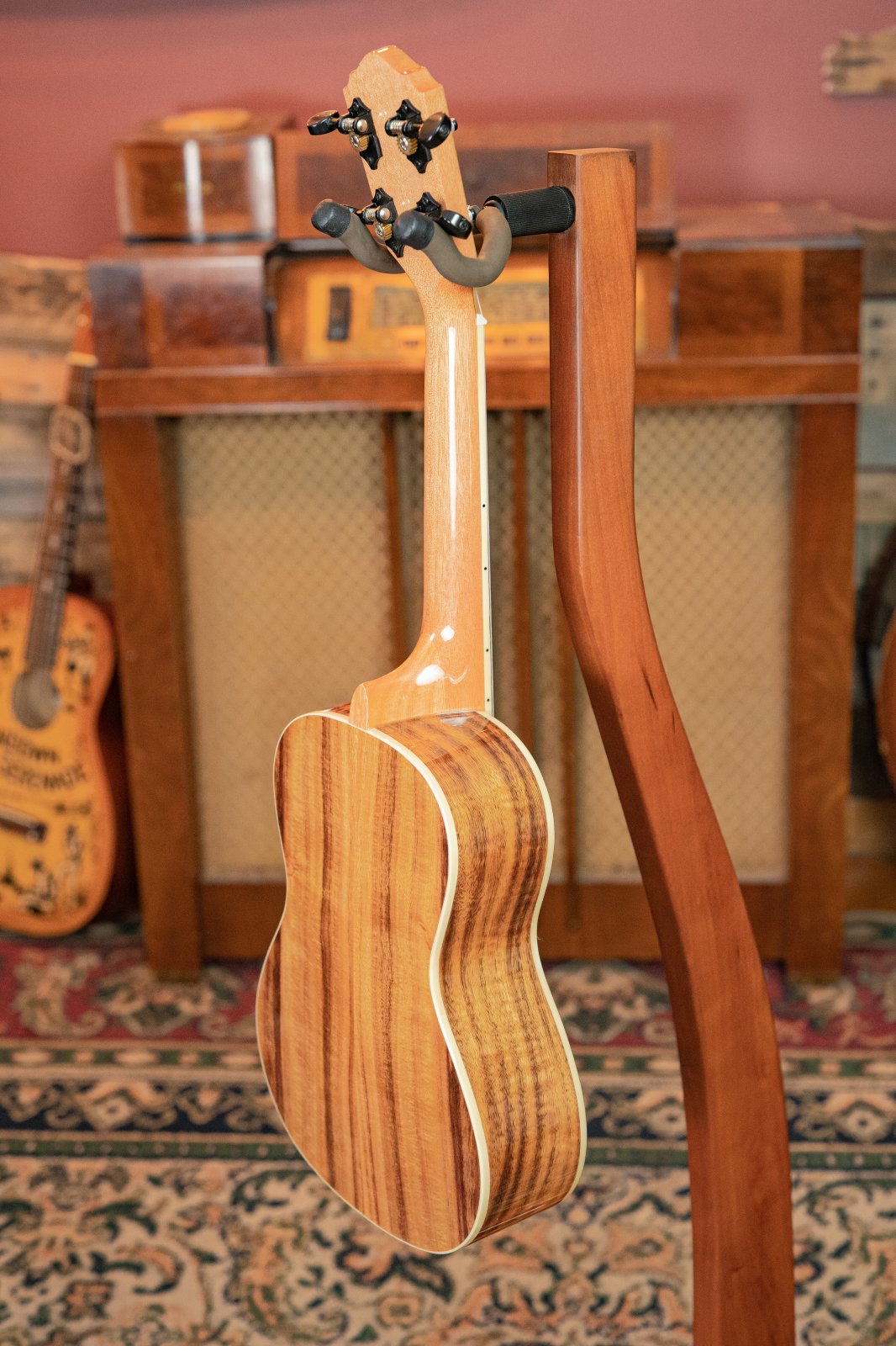 The Oscar Schmidt OU5K-A-U KOA Professional Concert Ukulele by Oscar Schmidt sits on a stand in a vintage-themed room, with an old guitar, radio, and patterned rug in the background.