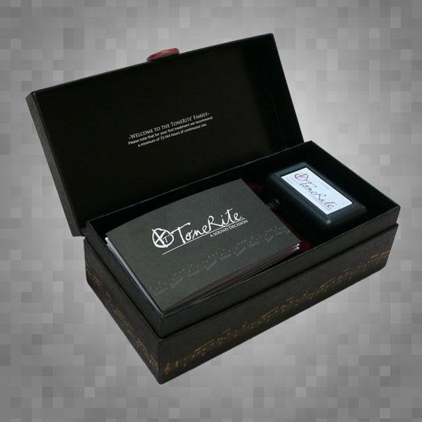 A black box with a hinged lid is open, revealing a ToneRite Guitar device and booklet from The Acoustic Shoppe—reflecting the detailed play-in process of vintage instruments. The box sits on a gray, pixelated background.