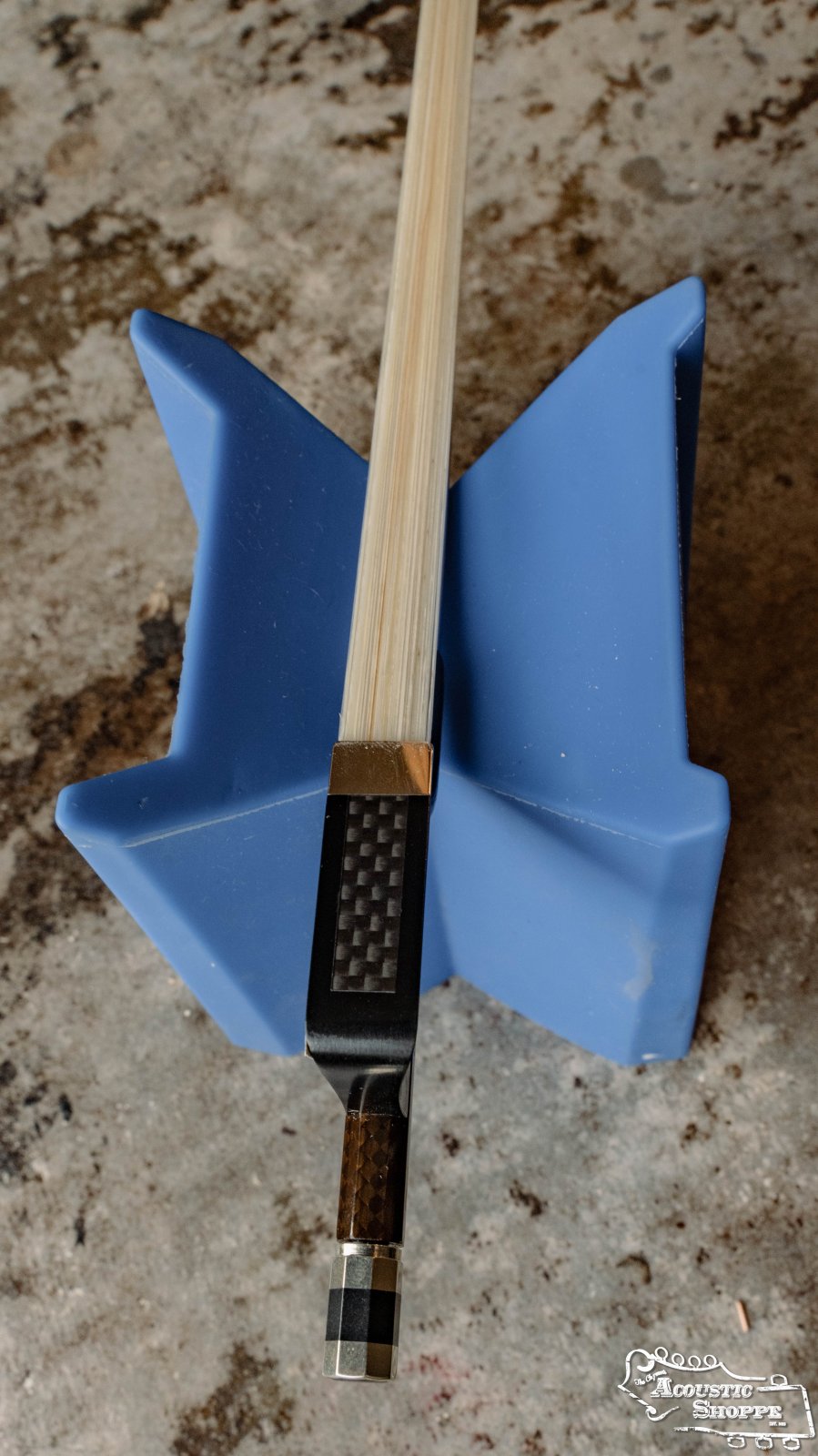 A CodaBow Luma Bow 4/4 Violin Bow by Coda Bow rests on a blue X-shaped holder atop a textured gray surface, showing both frog and tip with its stretched hair along the light carbon fiber stick.