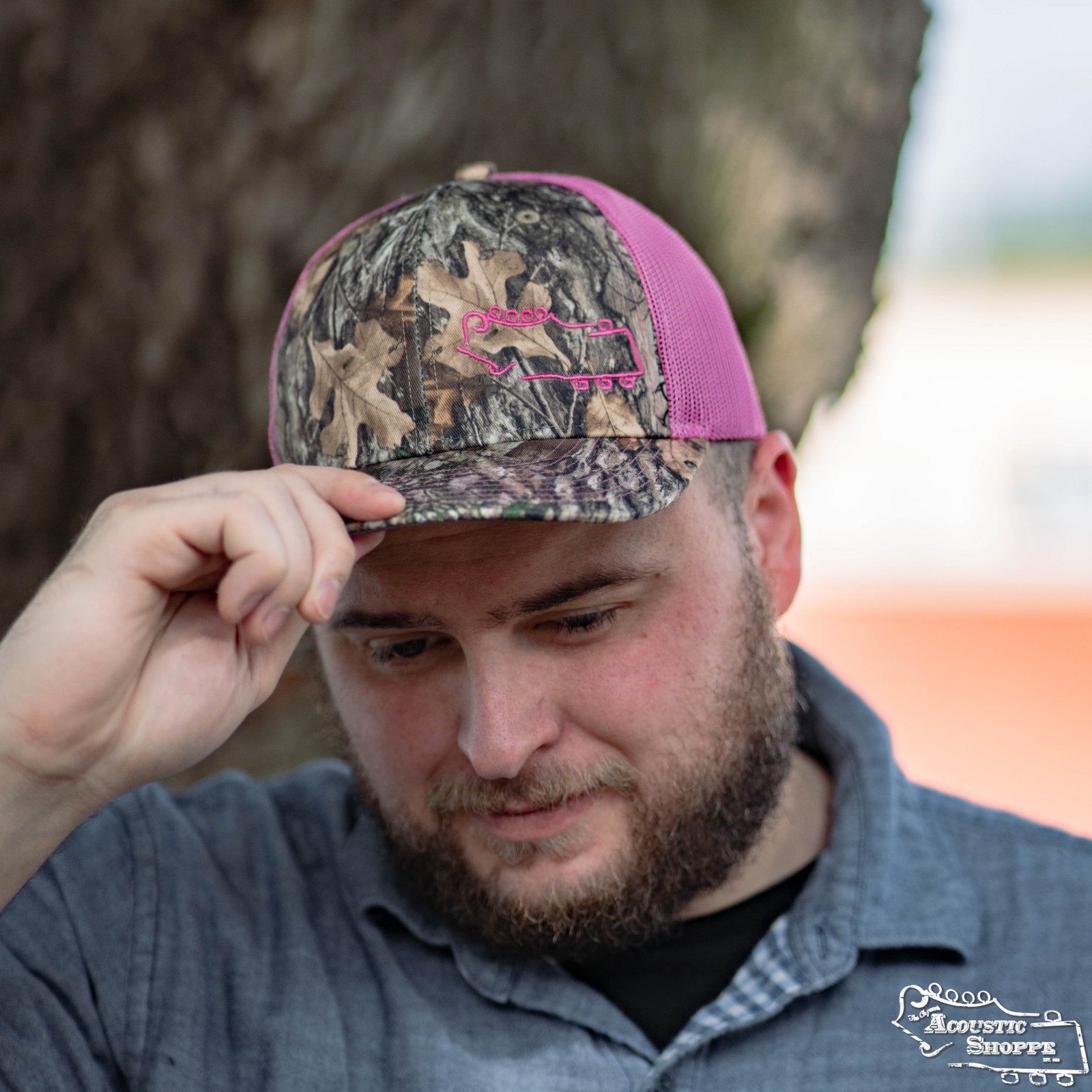 A bearded man stands outdoors by a tree, wearing an Amplify Screen Printing & Promotions Acoustic Shoppe Small Logo Outline Trucker Cap in neon pink with a Realtree Edge Camo bill, looking down as he holds the brim.