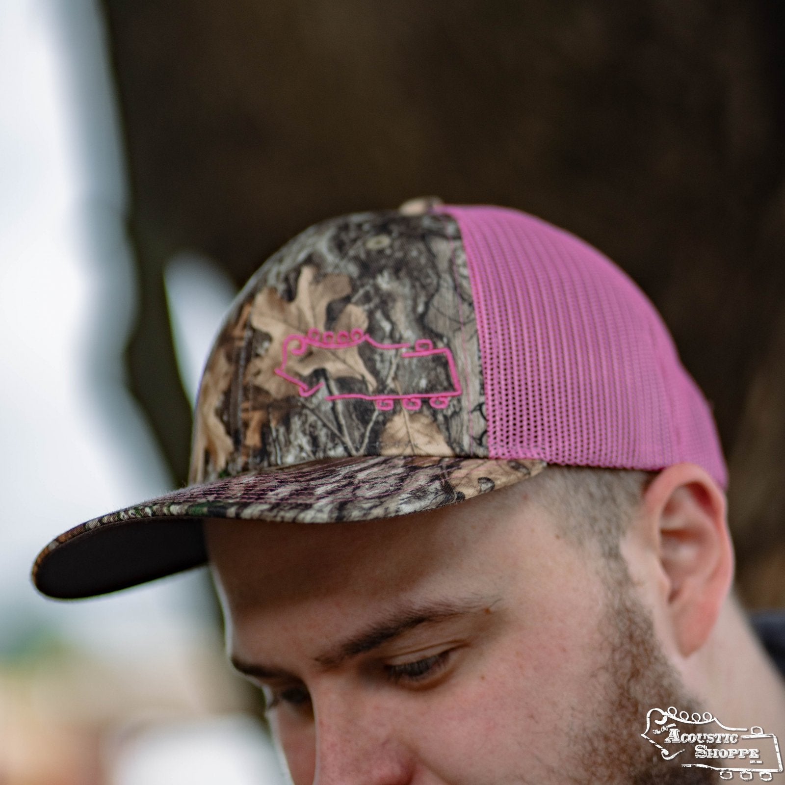A person wears the Acoustic Shoppe Small Logo Outline Trucker Cap in Neon Pink/Realtree Edge Camo by Amplify Screen Printing & Promotions, featuring a pink chainsaw logo—ideal for unique headwear fans. Photo is outdoors and hat-focused.