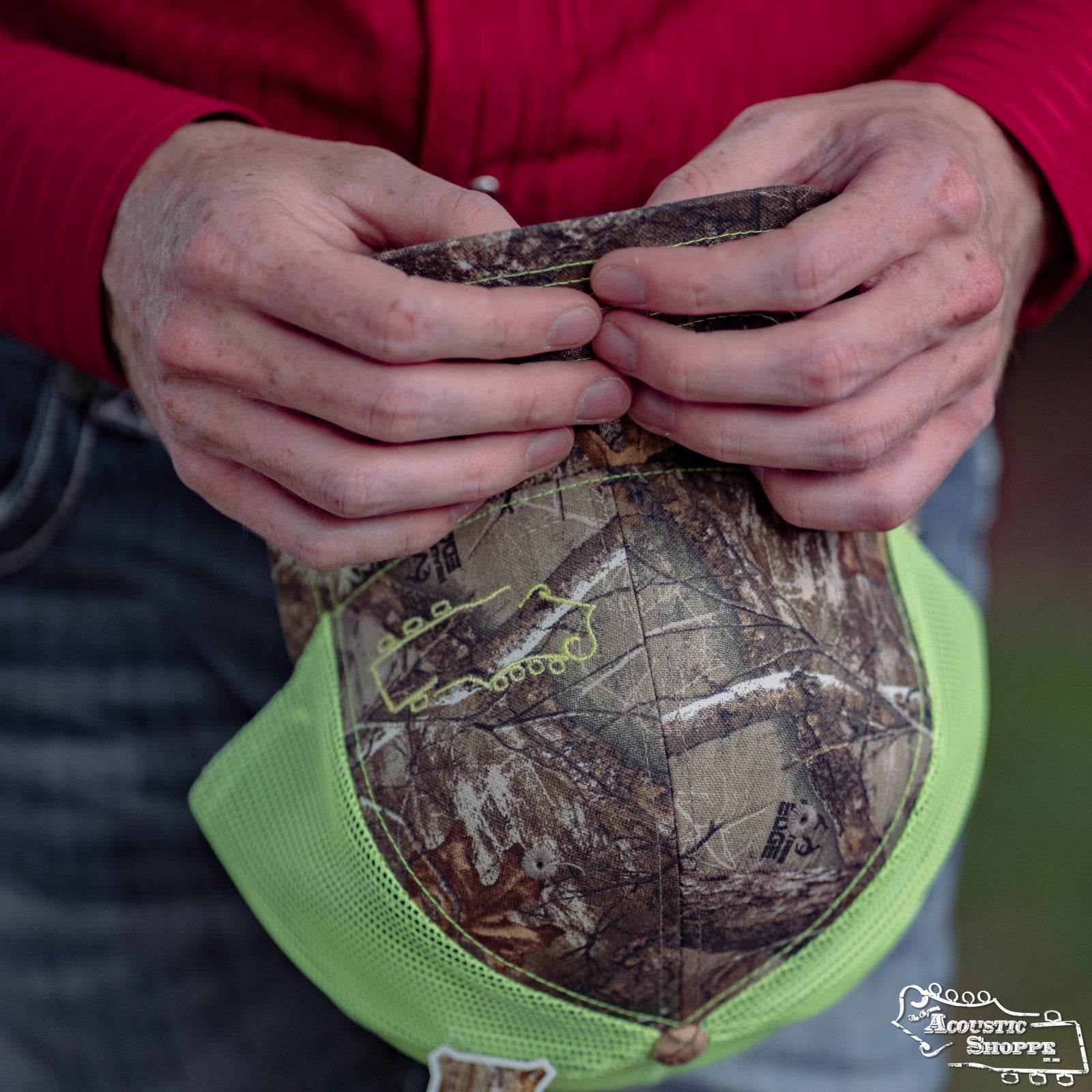 A person outdoors in a red shirt holds the Amplify Screen Printing & Promotions Acoustic Shoppe Small Logo Outline Trucker Cap in Neon Yellow / Realtree Edge Camo, focusing on its adjustable strap at the back.