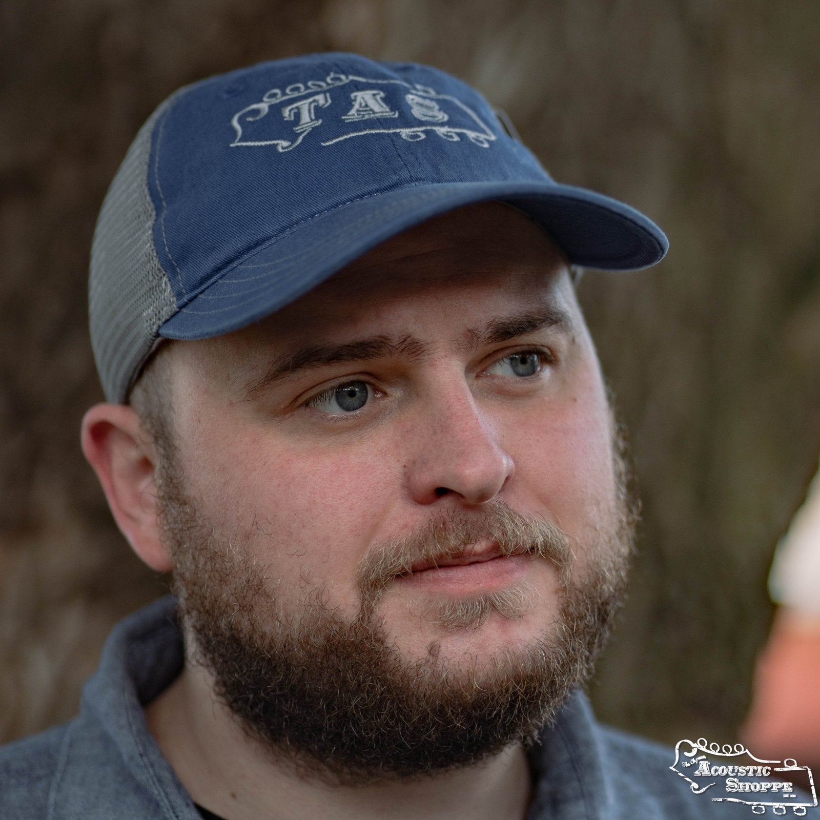 A bearded man in a gray shirt and Acoustic Shoppe Large TAS Logo Trucker Cap (Royal/Charcoal) by Amplify Screen Printing & Promotions stands outdoors, gazing slightly to the side in front of a tree trunk.