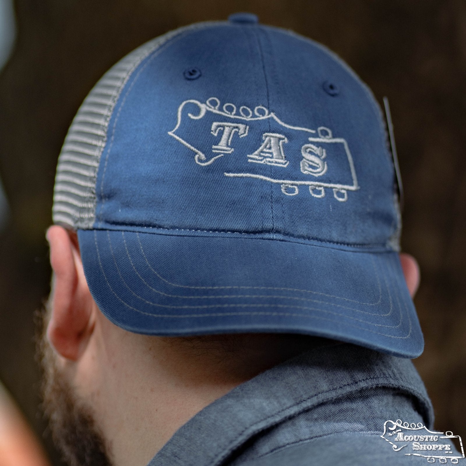 A person faces away from the camera wearing the Acoustic Shoppe Large TAS Logo Trucker Cap in Royal/Charcoal by Amplify Screen Printing & Promotions, featuring a mesh back, guitar headstock design, and TAS embroidered on the front.