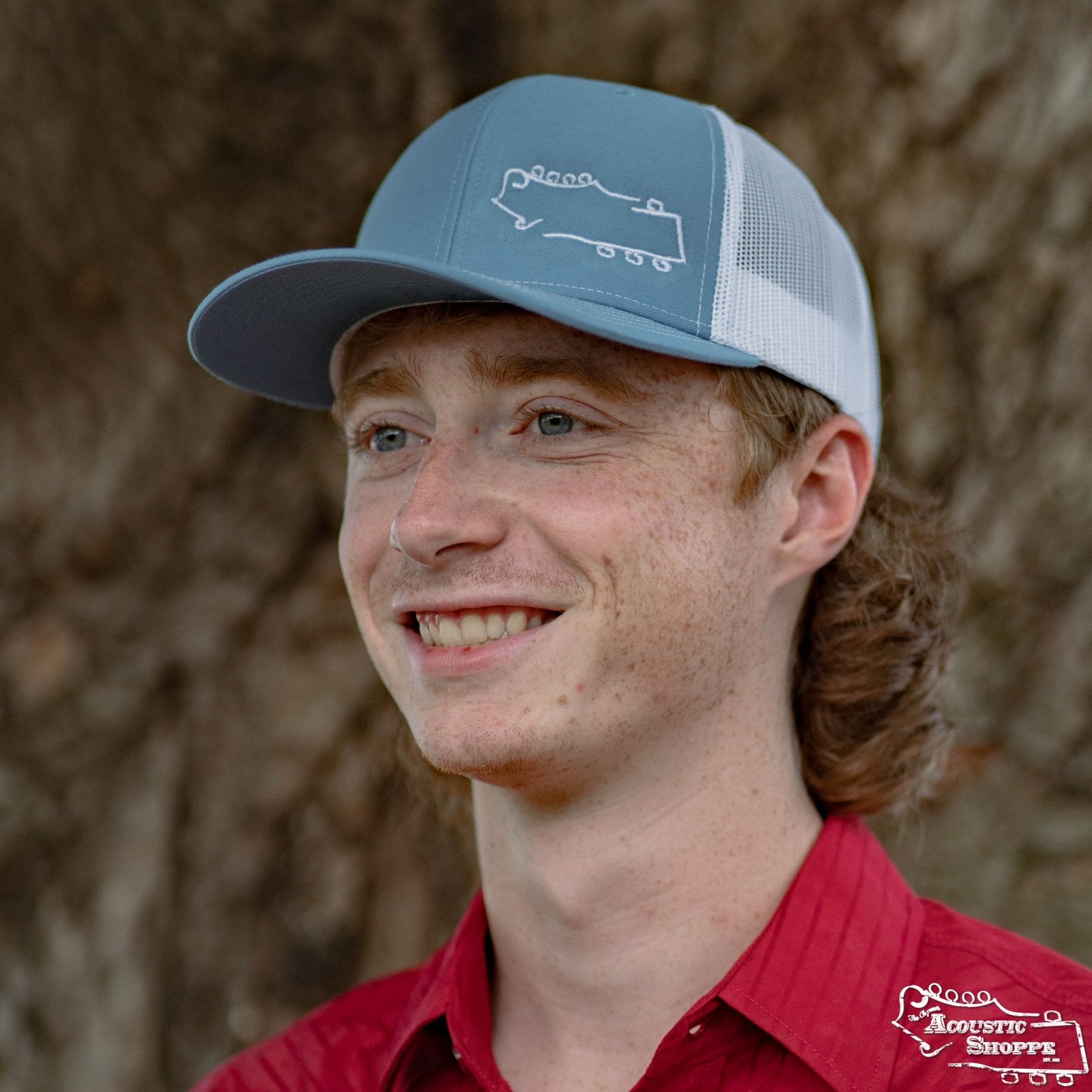 A young man with fair skin, blue eyes, and red hair is smiling in front of a tree trunk while wearing an Amplify Screen Printing & Promotions Acoustic Shoppe Small Logo Outline Trucker Cap in blue and white and a red collared shirt.