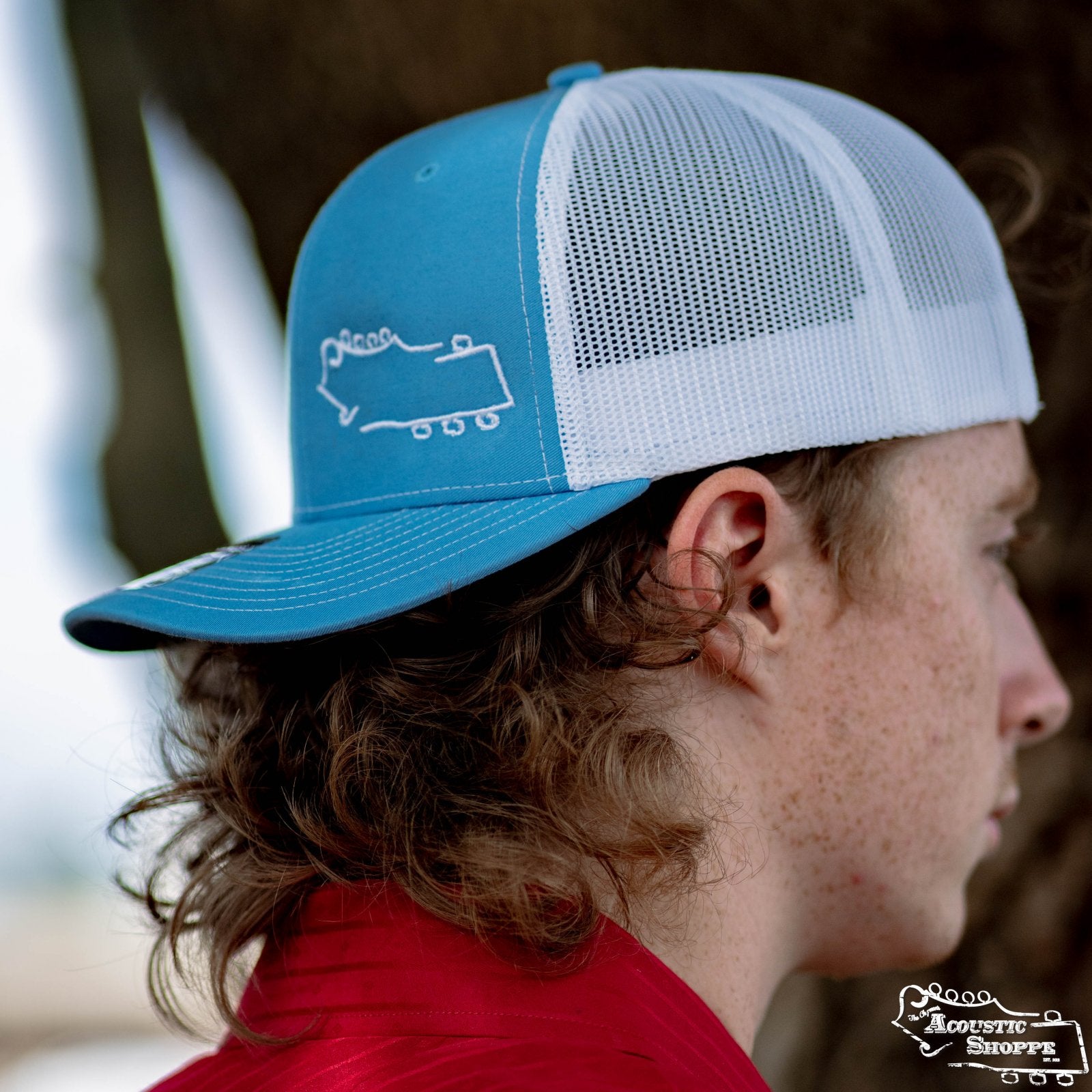 A person with curly hair wears the Acoustic Shoppe Small Logo Outline Trucker Cap in blue and white by Amplify Screen Printing & Promotions, paired with a red shirt. The side profile is shown against a blurred background.
