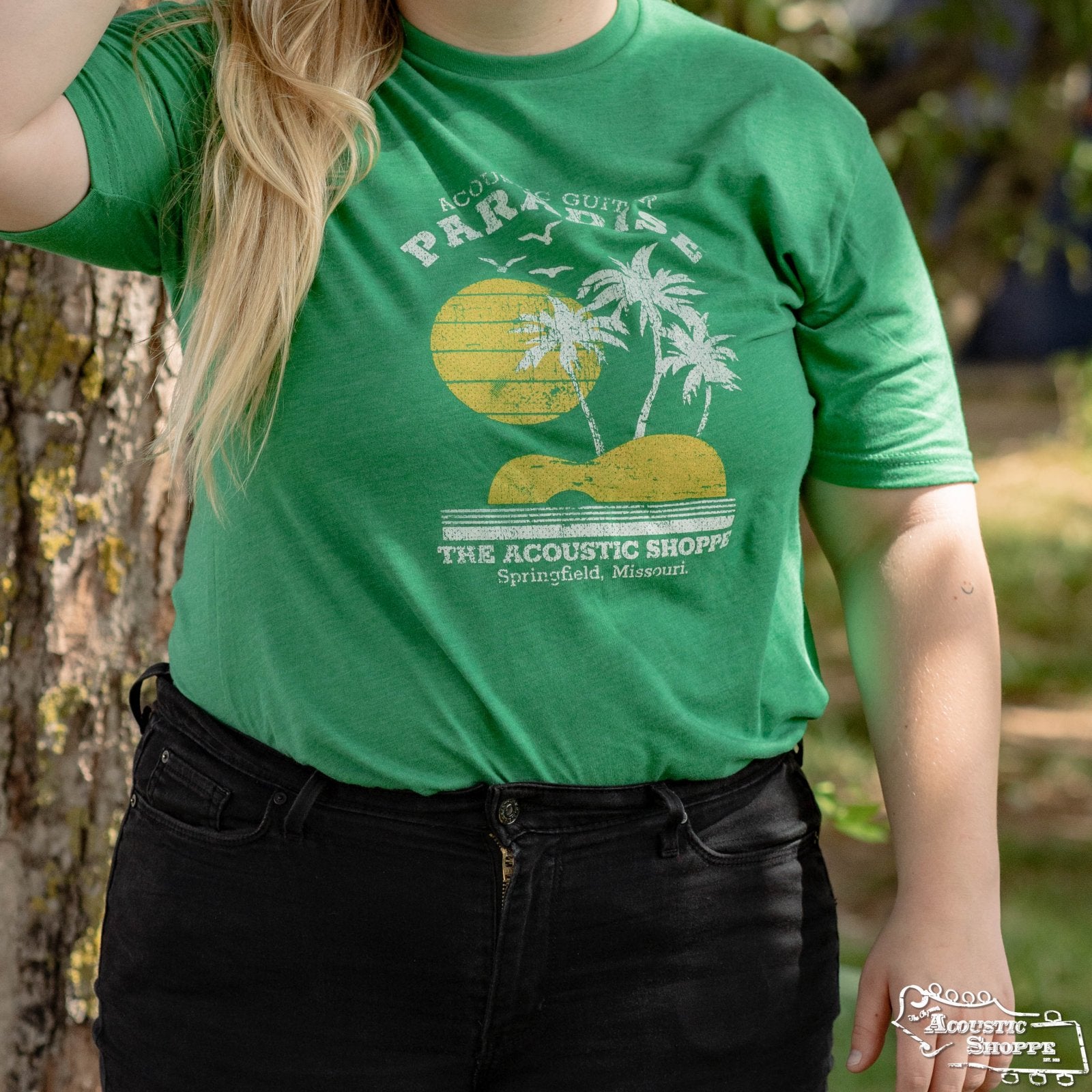 A person wearing The Acoustic Shoppe Paradise Design T-Shirt (Green) by Amplify Screen Printing & Promotions, featuring palm trees, sun, and Acoustic Paradise text, stands outdoors near a tree in Springfield, Missouri.