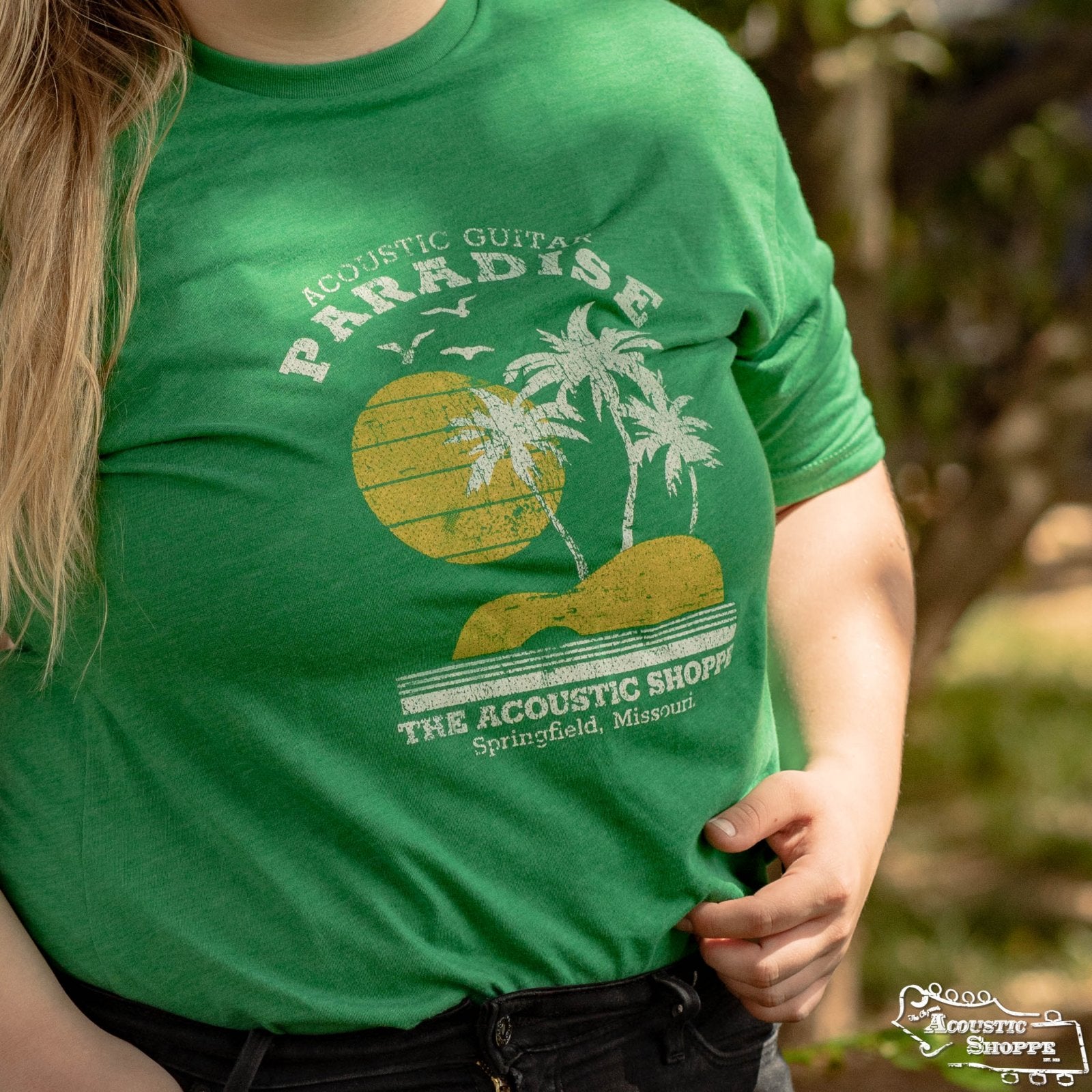 Wearing the green Paradise Design T-Shirt by Amplify Screen Printing & Promotions, featuring palm trees, sun, and “Acoustic Guitar Paradise, The Acoustic Shoppe, Springfield, Missouri”—a must-have for acoustic music fans.