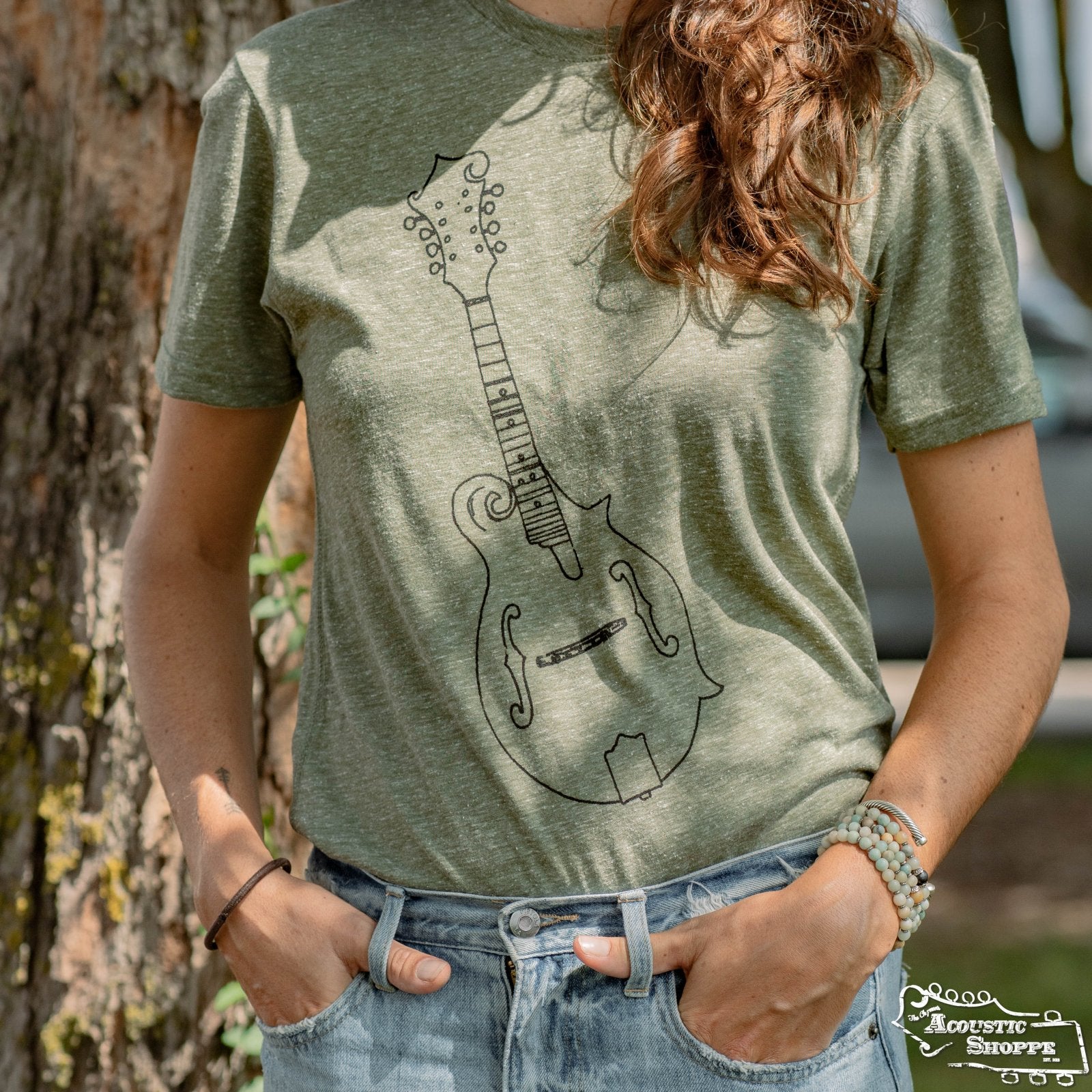 A woman stands outdoors among trees wearing the Amplify Screen Printing & Promotions TAS Hand-Drawn Mandolin Heathered Green T-Shirt, featuring a simple black mandolin illustration. She poses with one hand in her pocket and the other on her hip.