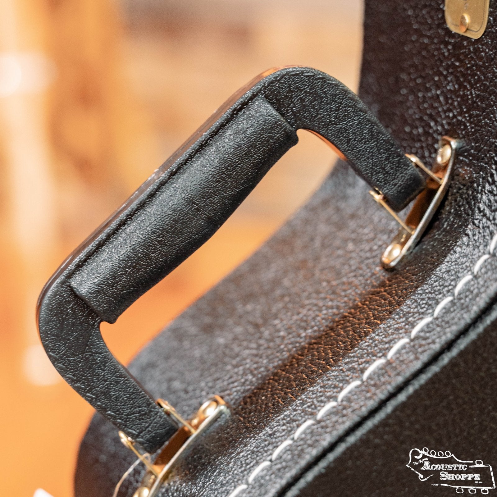 Close-up of a black leather handle with white stitching and gold hardware on the TKL 7820 Premier Jumbo 6/12 String Hardshell Guitar Case. The Acoustic Shoppe logo appears in the bottom right corner.