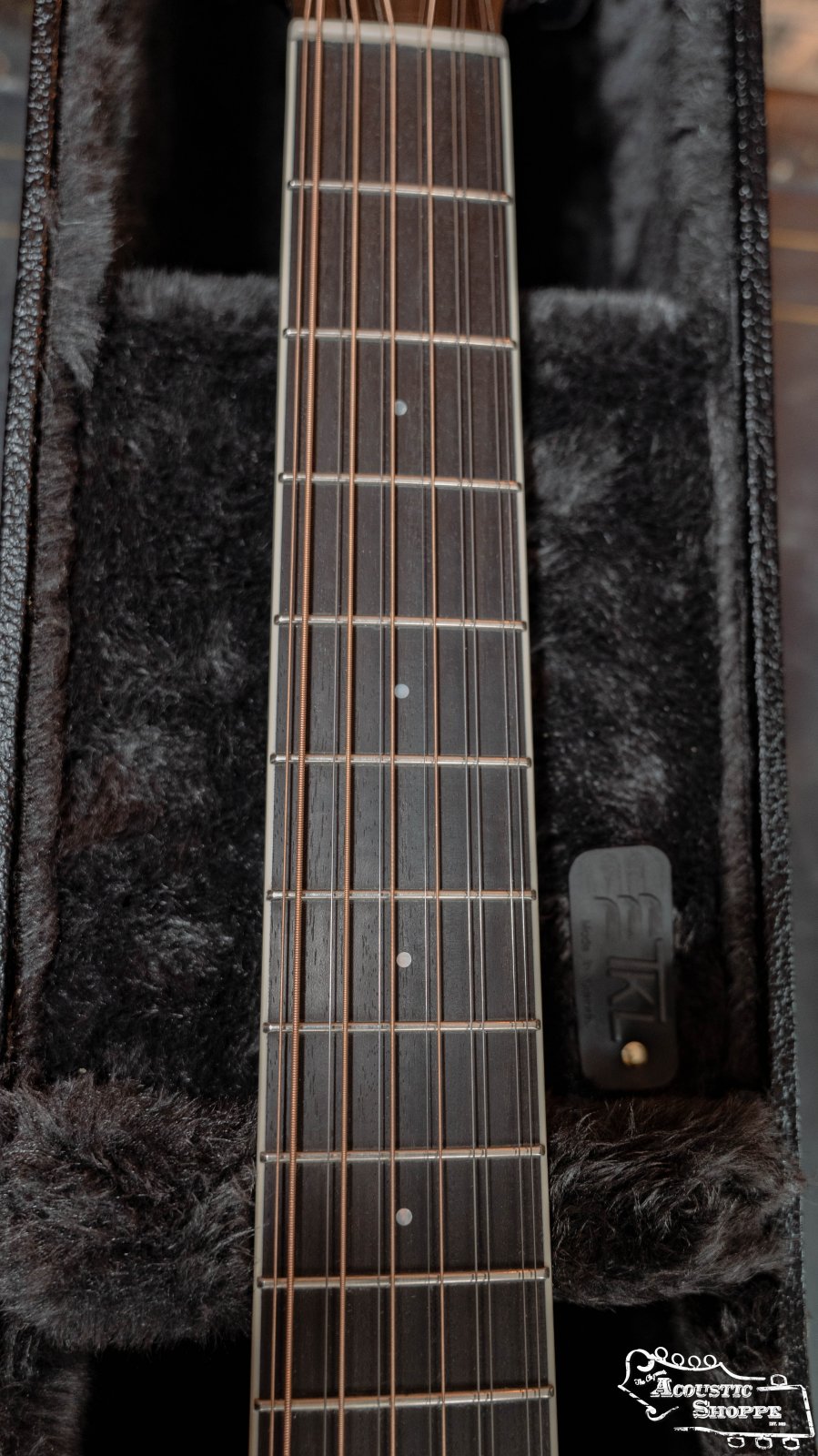Close-up of a guitar neck with frets and dot inlays resting inside a plush-lined TKL 7820 Premier Jumbo 6/12 String Hardshell Guitar Case by TKL, with a small black device attached near the bottom right corner of the case.