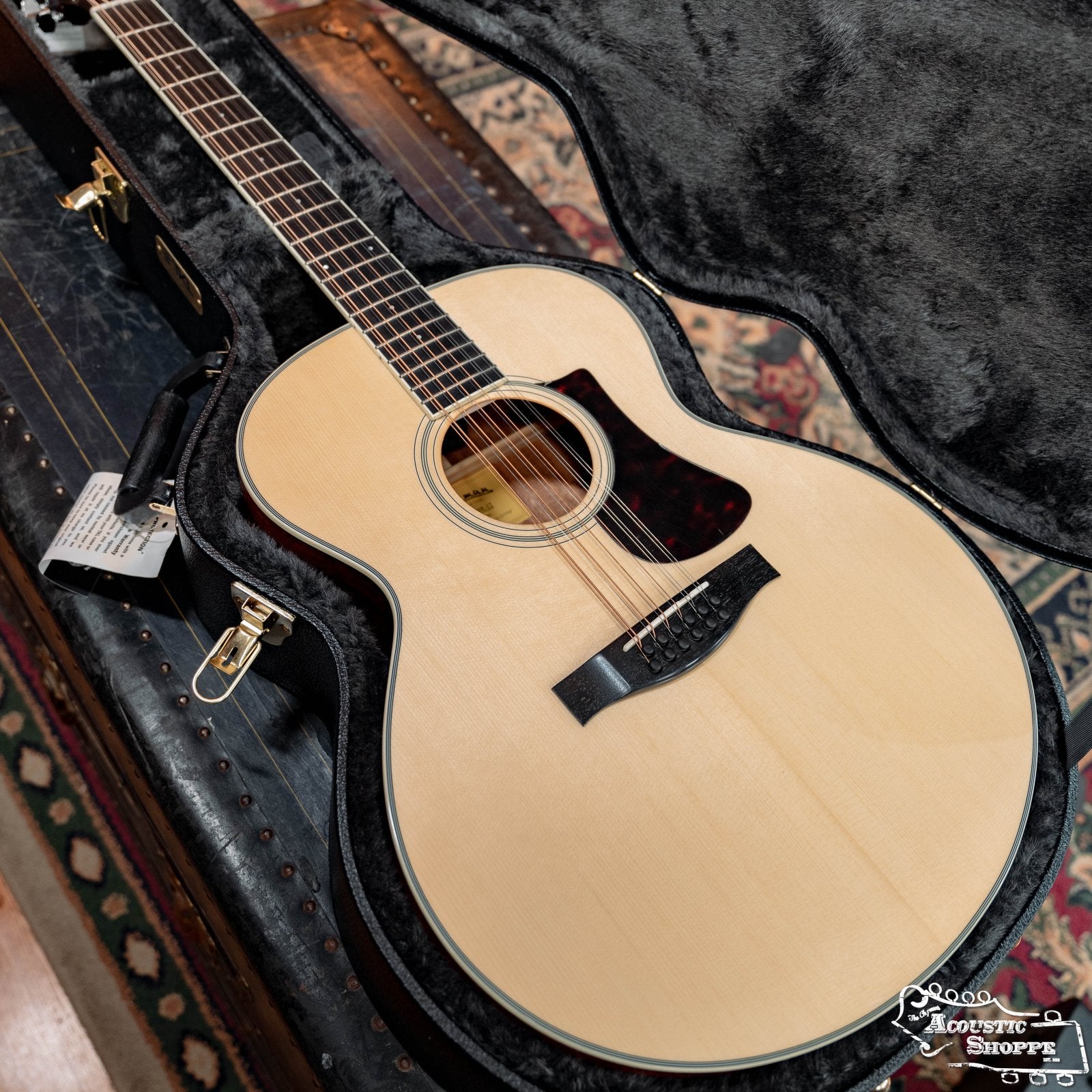 A light-colored acoustic guitar with a black pickguard sits in an open TKL 7820 Premier Jumbo 6/12 String Hardshell Guitar Case, lined with plush black and resting on a patterned rug atop a vintage trunk.