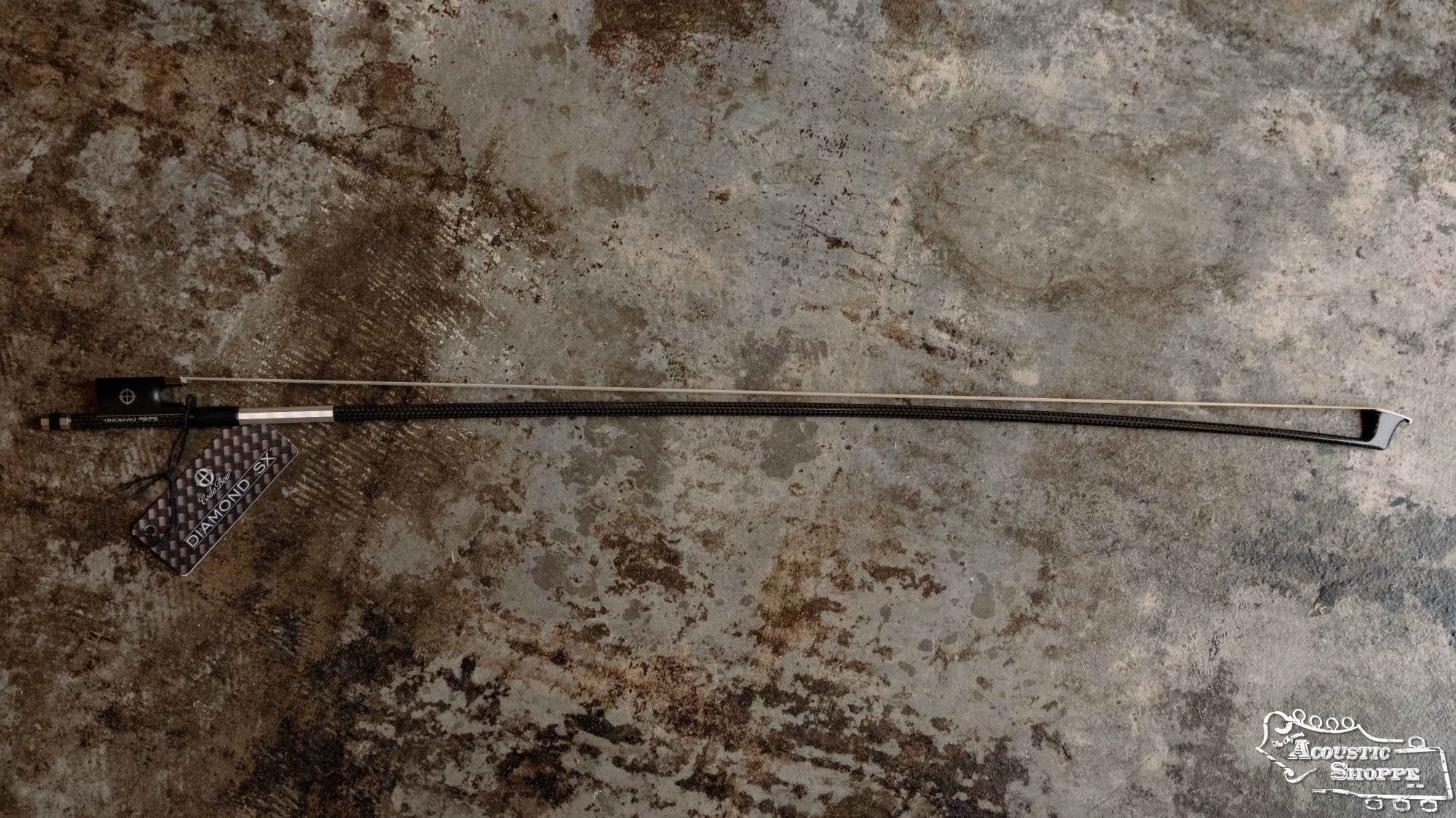 A Coda Bow Diamond SX 4/4 carbon fiber violin bow with tag rests on a mottled brown-gray surface, ideal for gigging musicians. The Acoustic Shoppe logo is displayed in the bottom right corner.