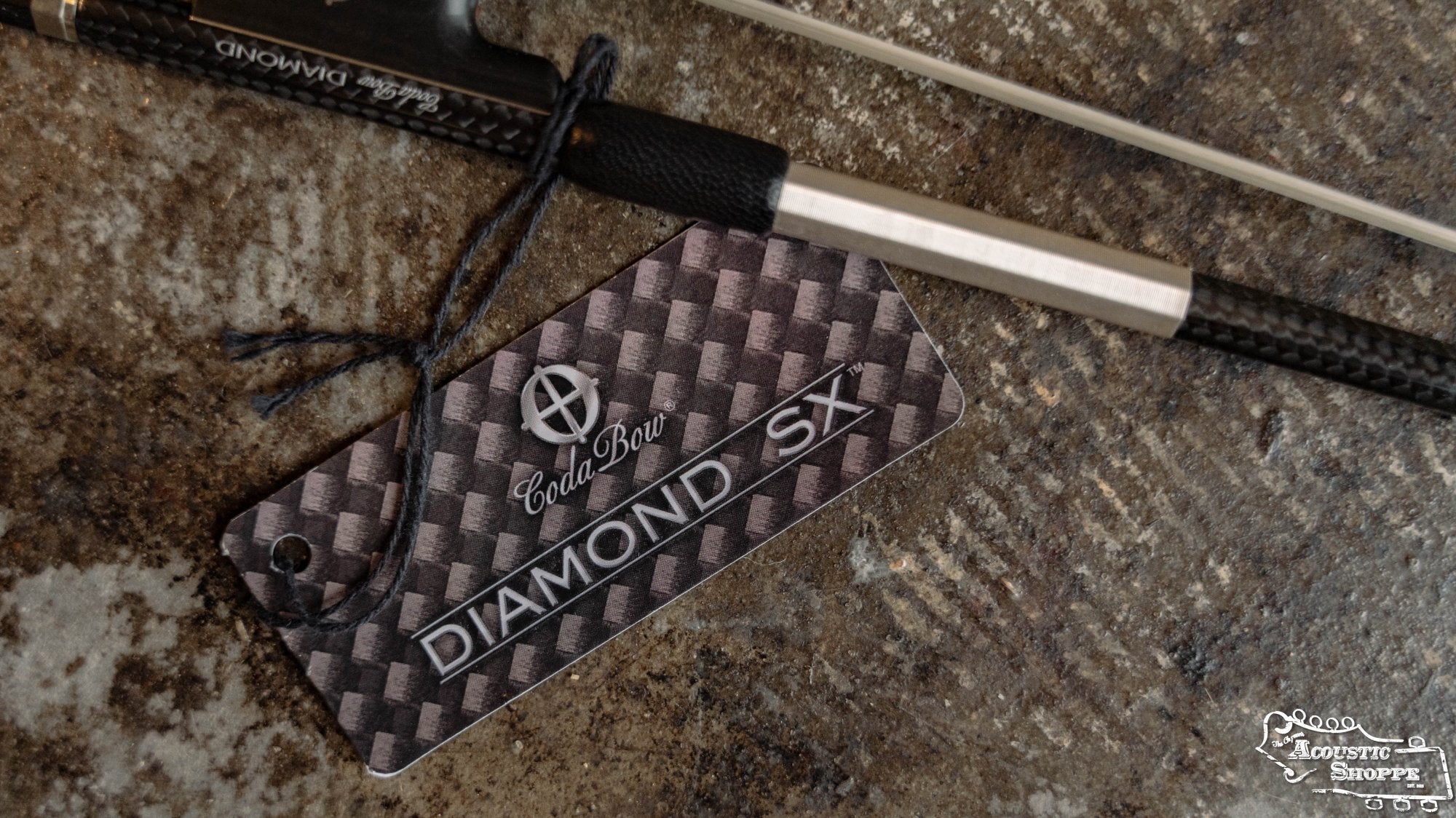 A Coda Bow Diamond SX 4/4 Violin Bow and its carbon fiber-patterned tag are displayed on a textured surface, with The Acoustic Shoppe logo visible in the lower right corner.